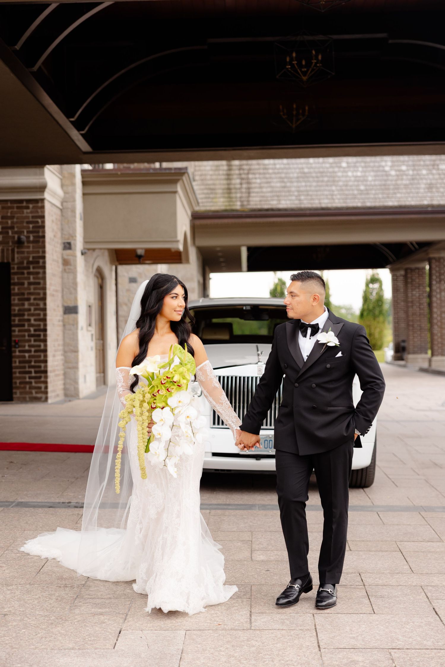 A bride in a white lace gown and groom in black tuxedo stand together holding hands in front of a classic white car.