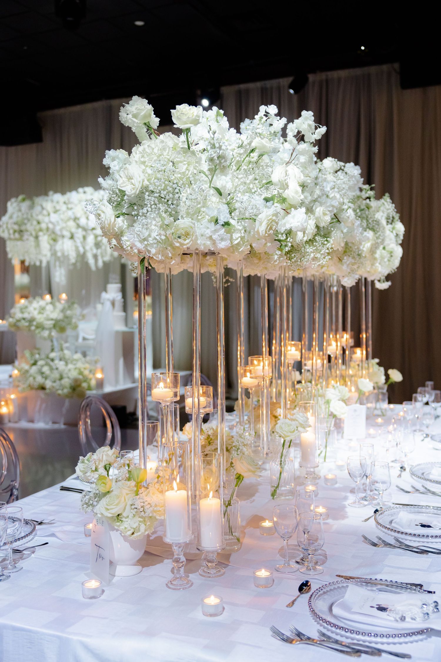 Elegant white floral centerpiece with tall glass stems and candles on a formal dining table at a wedding reception.