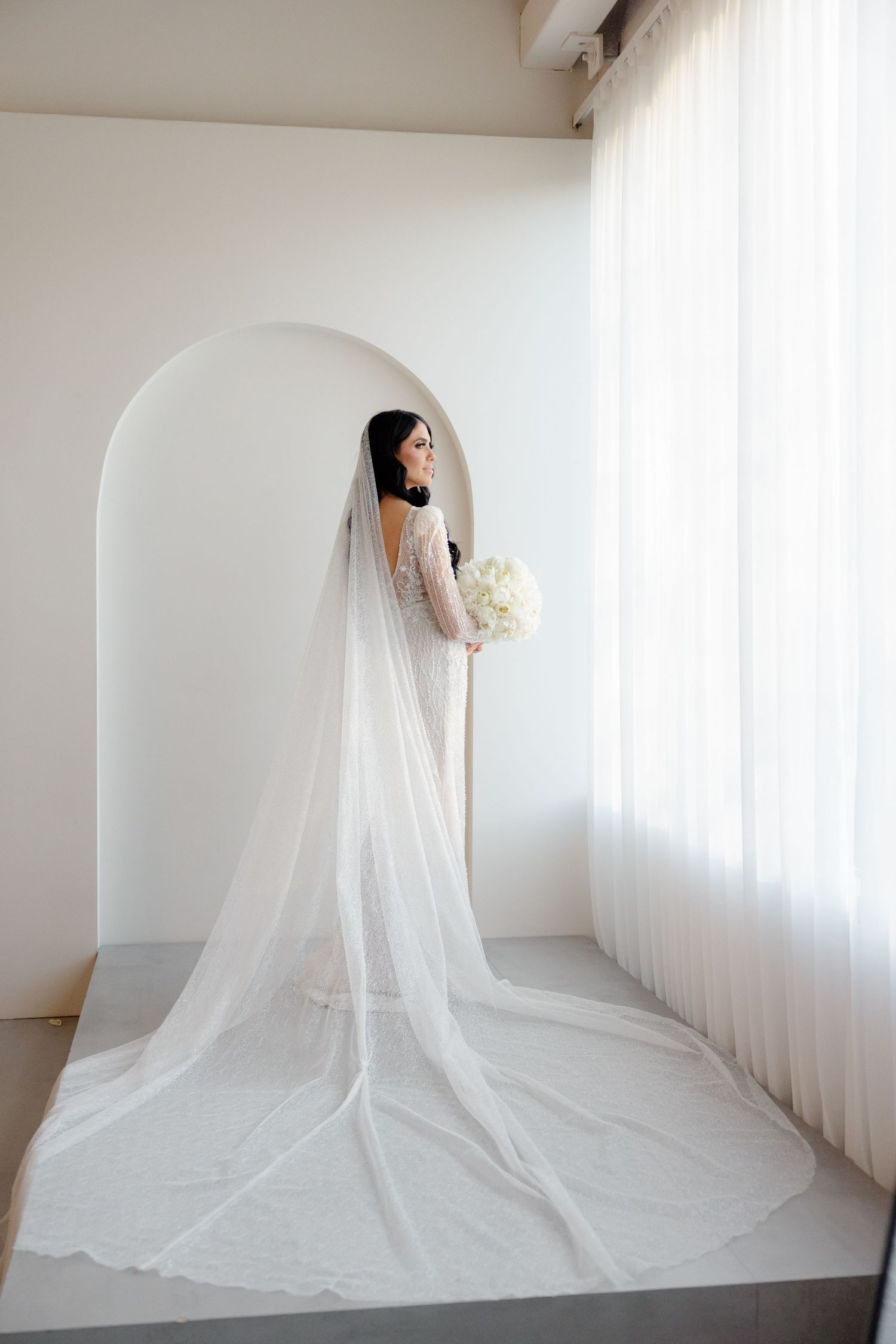 Bride in long sleeve lace gown with cathedral train poses against minimalist white backdrop.