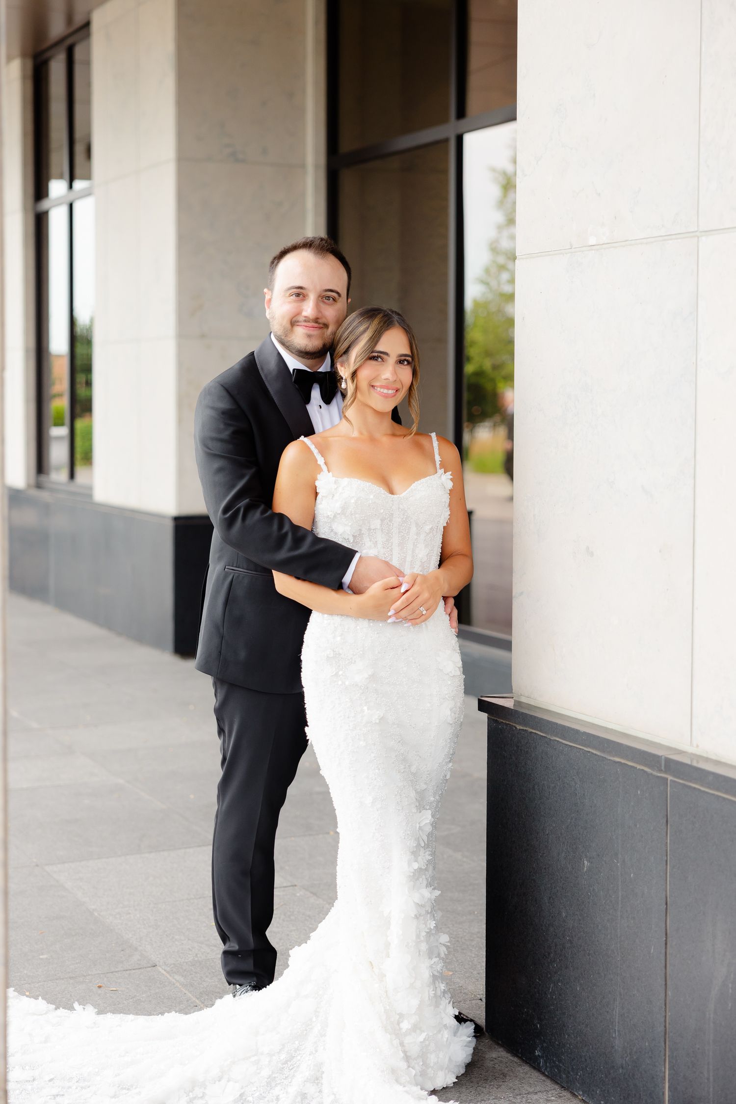 A couple dressed in wedding attire shares romantic moments against a white building exterior for their wedding photos.