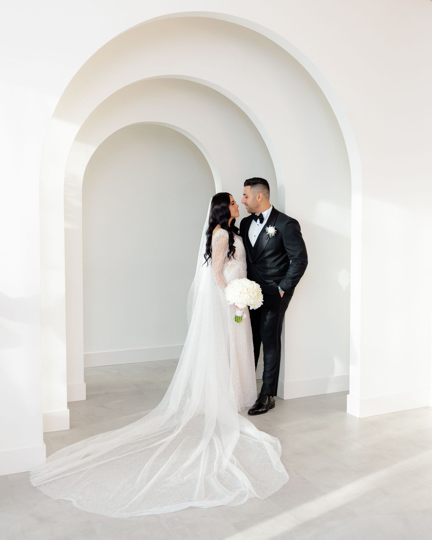 Elegant wedding couple stands under white archway with bride holding white bouquet and wearing flowing gown.
