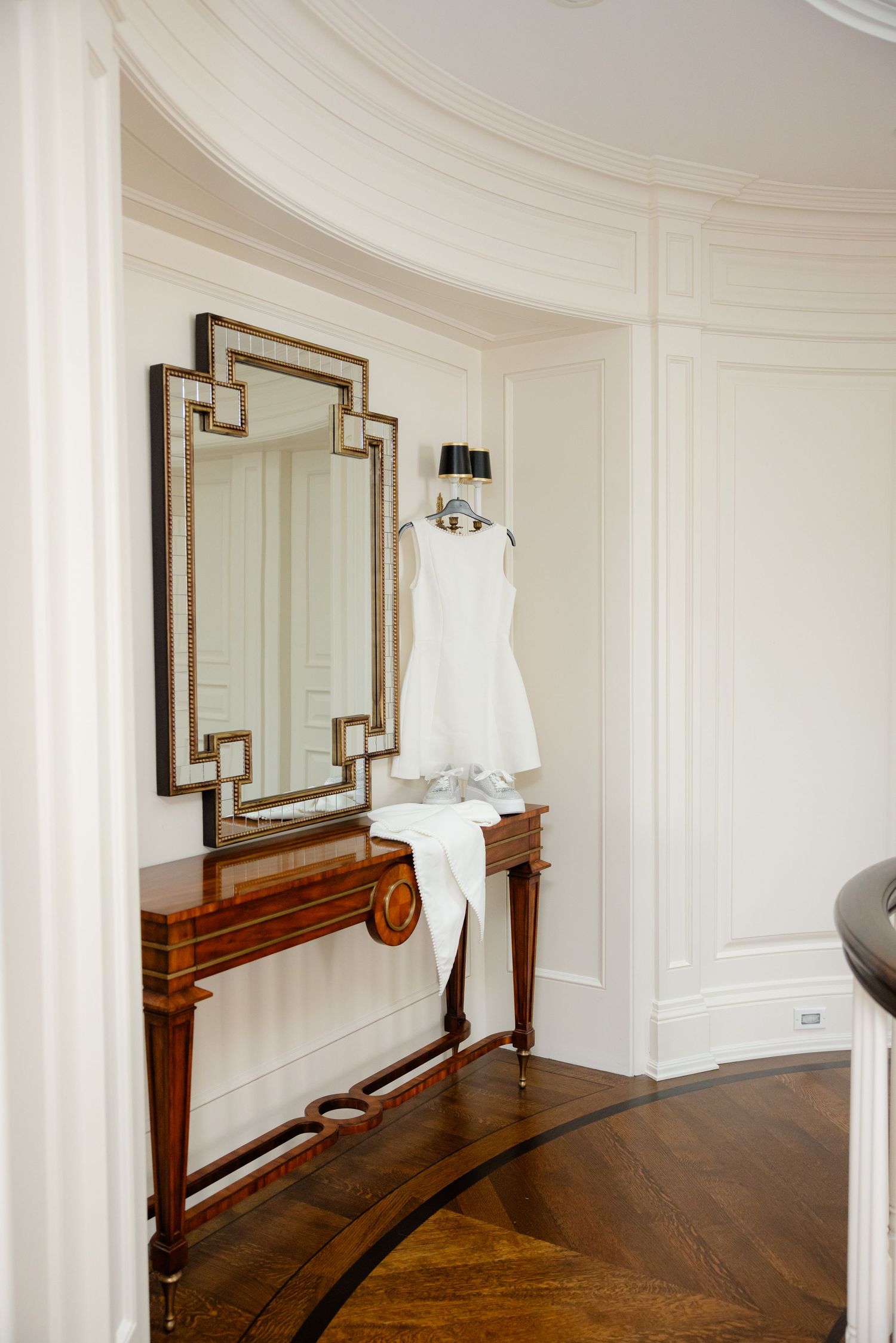 Elegant curved hallway with ornate mirror and wooden console table displaying white wedding attire.
