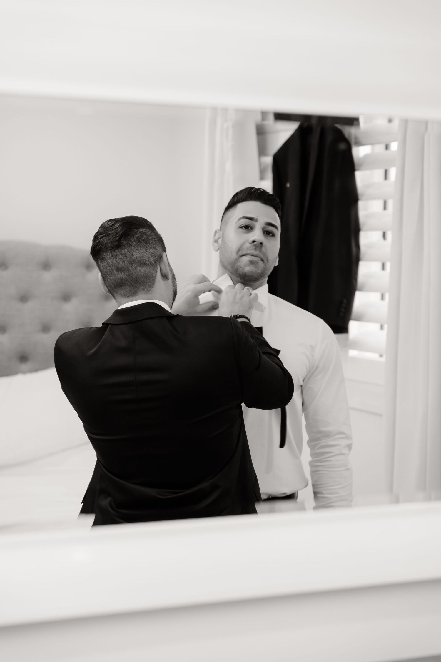 Black and white reflection of groomsmen helping with bow tie preparation in elegant mirror.