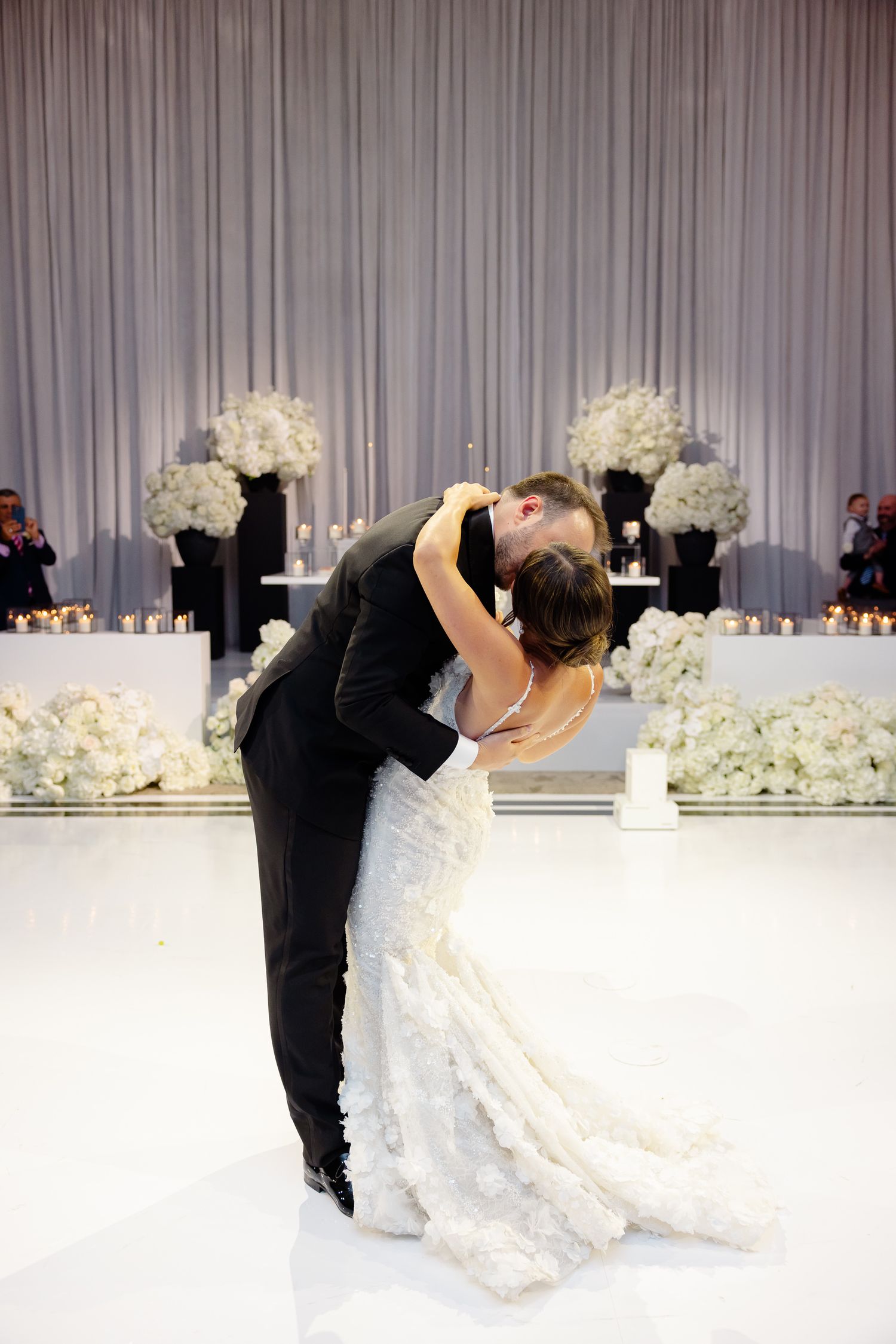 Romantic wedding reception scene with couple dancing on white floor surrounded by white floral arrangements and candles.