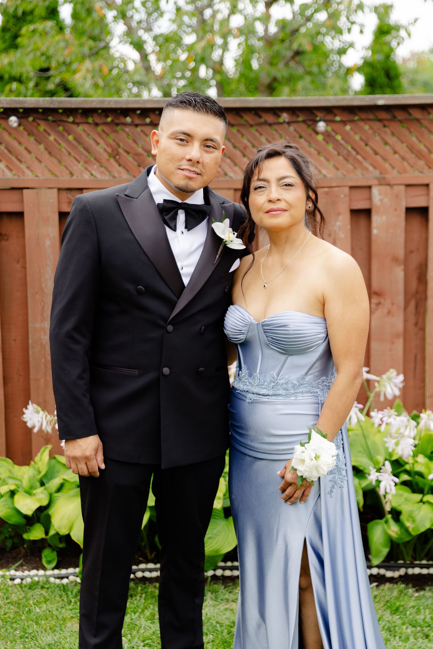 A wedding party in formal attire poses together outdoors against a wooden fence and garden backdrop.
