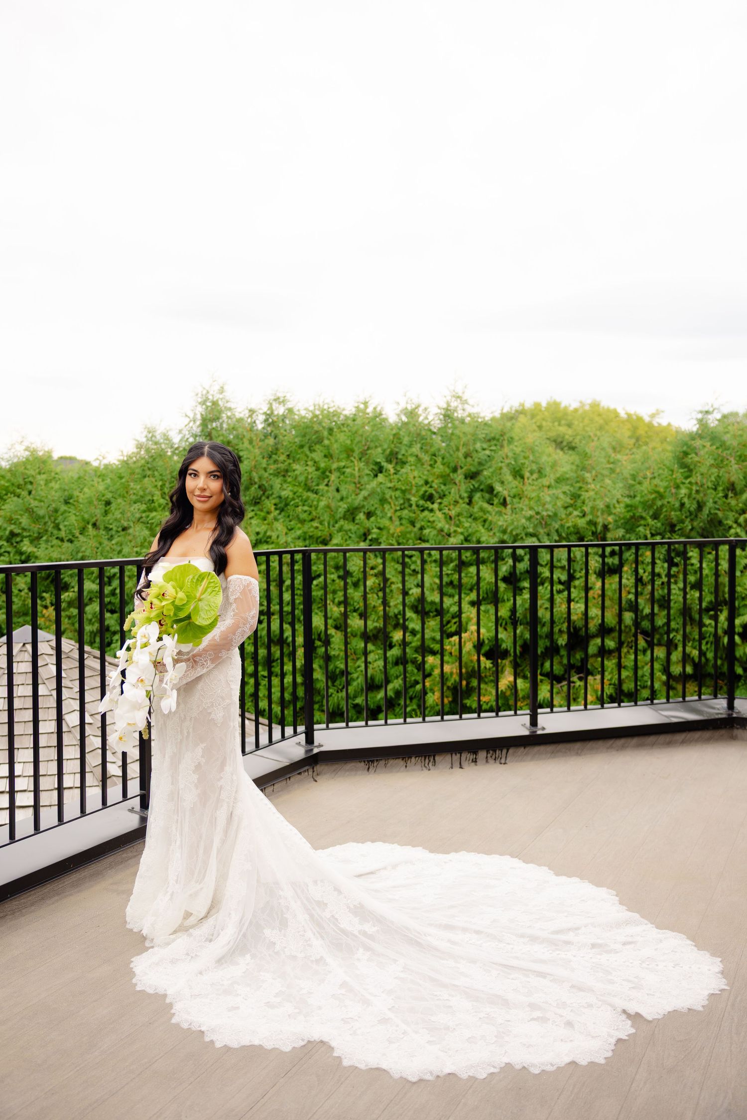 A bride in a flowing lace wedding gown poses gracefully on a balcony overlooking lush greenery.