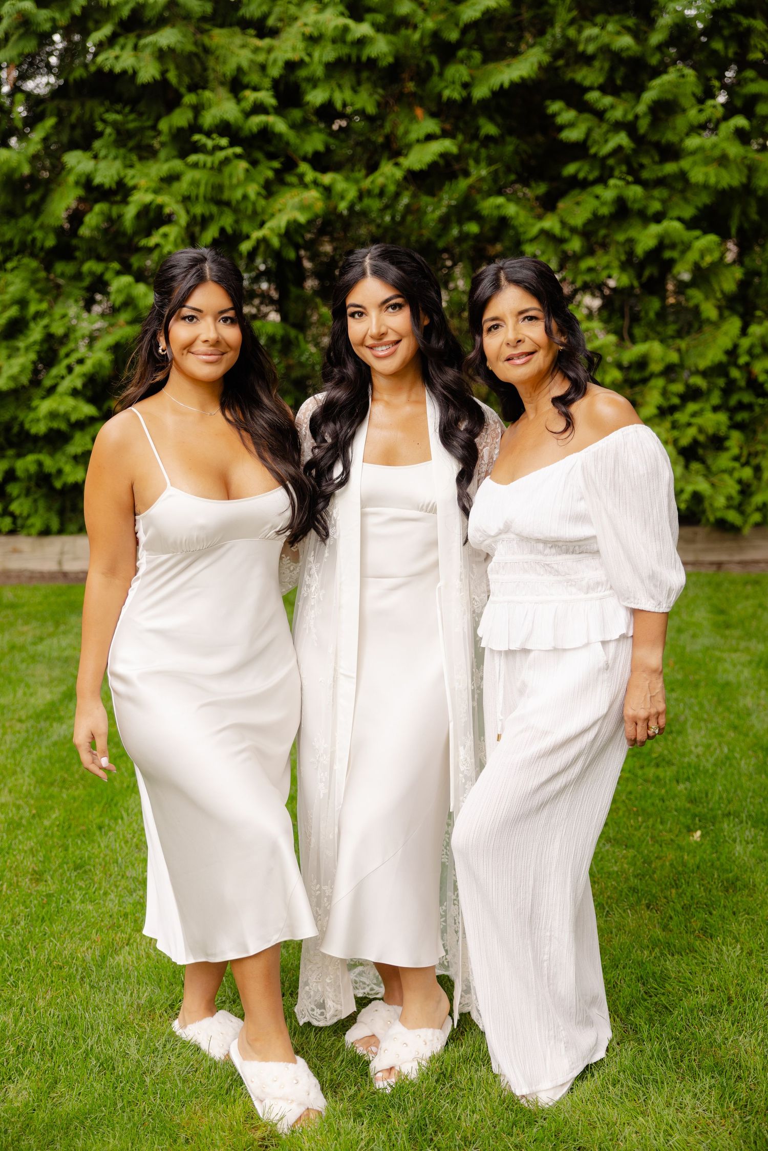 Three people in white dresses posing together outdoors on grass with lush greenery behind them.