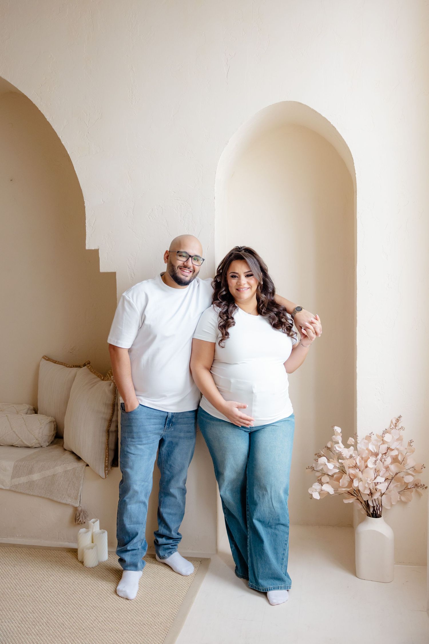 A couple in matching white t-shirts and jeans stand together in front of an arched wall with minimalist decor.