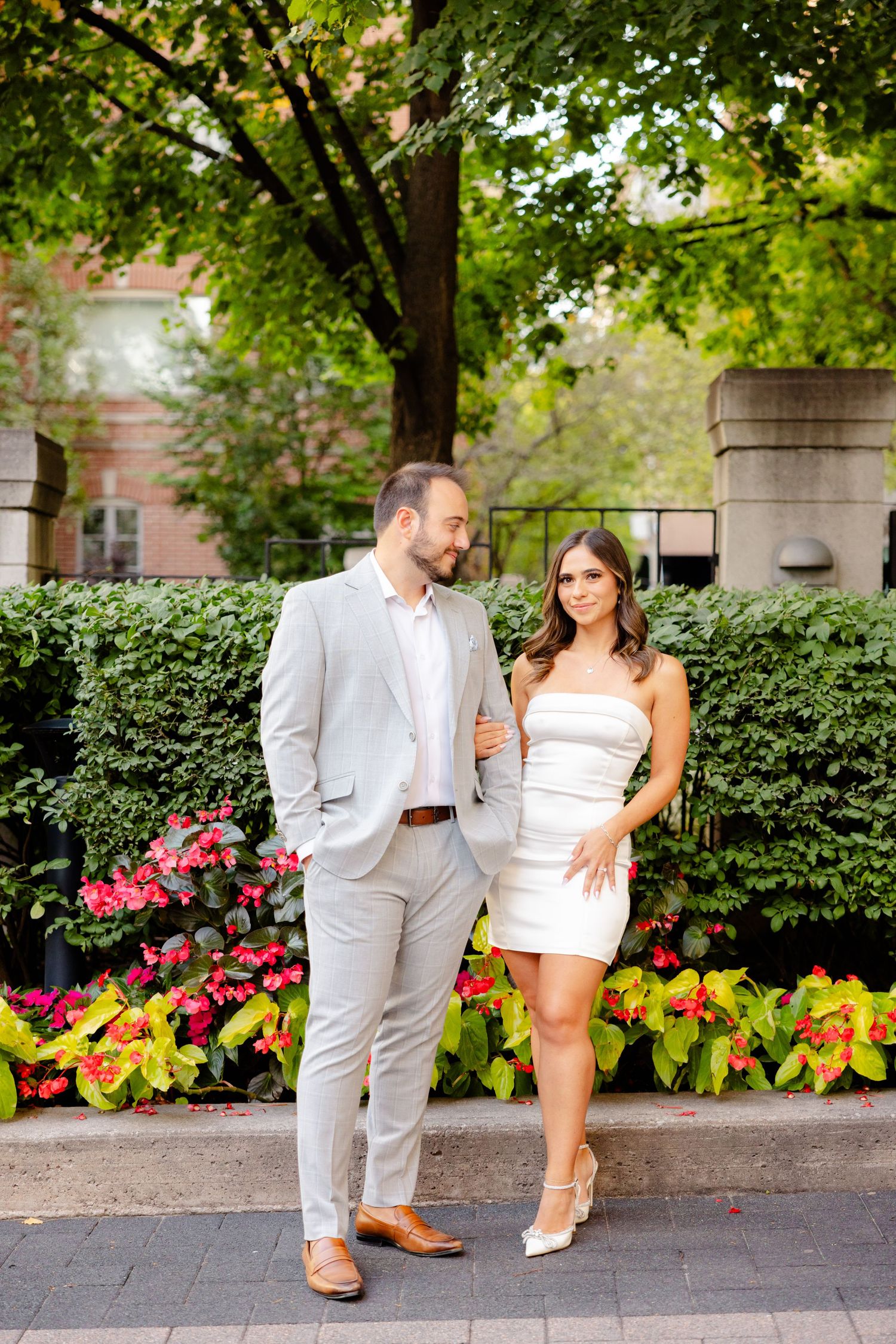 A fashionable couple in light colored formal wear poses against a backdrop of greenery and flowers.