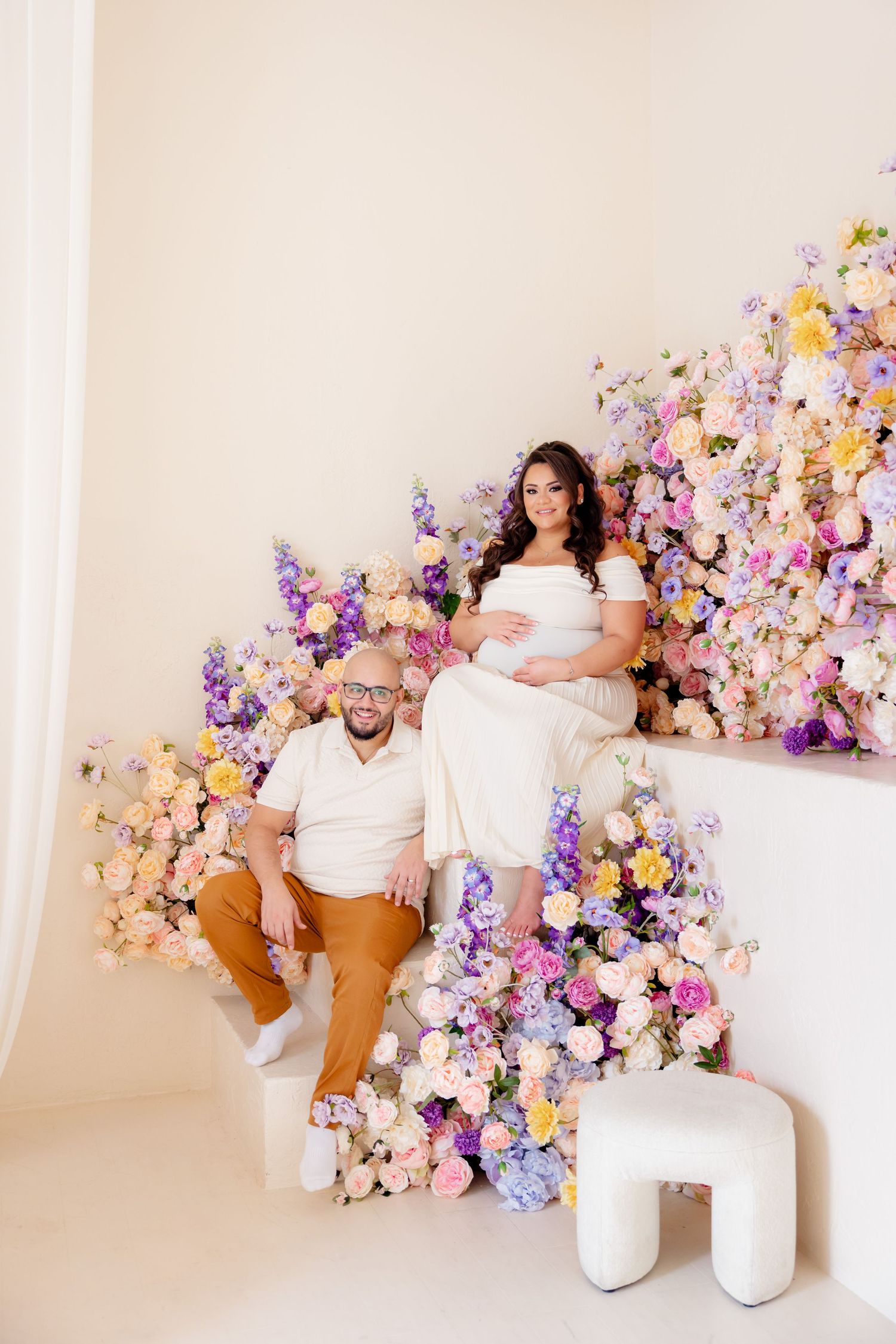 A couple poses in a romantic studio setting with elegant floral arrangements in purple, pink and white tones.