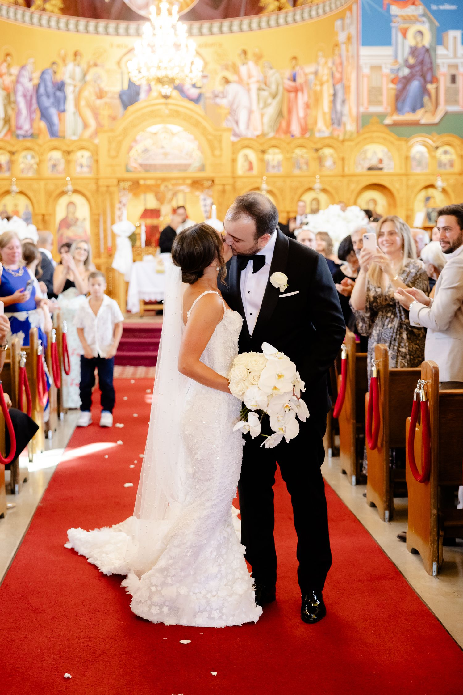 Newlyweds share a romantic kiss on the red carpeted aisle of an ornate Orthodox church with golden iconography.