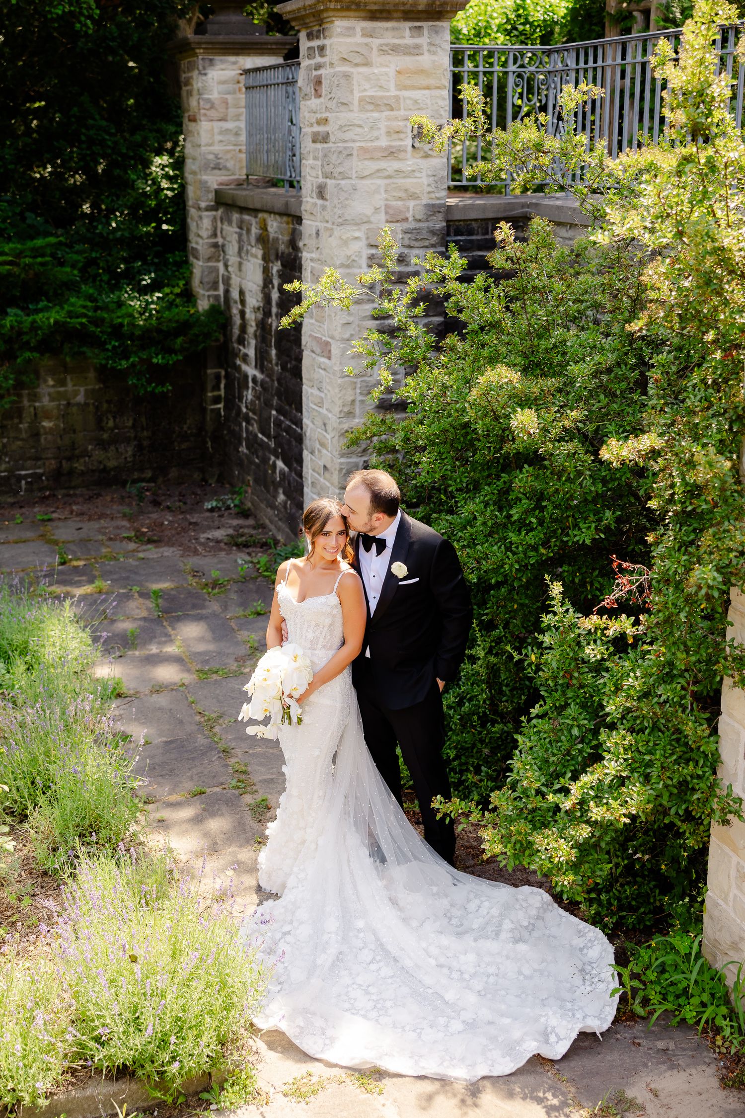 Elegant bride and groom share an intimate moment in a picturesque garden with stone columns and flowering plants.