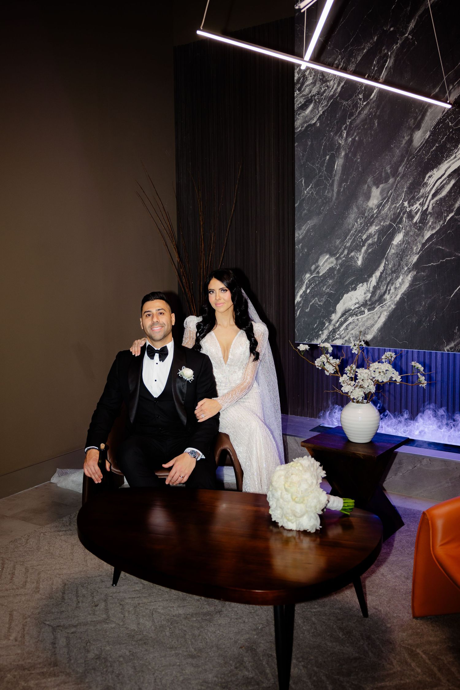 Elegant wedding couple poses near modern decor with marble wall and floral arrangements.