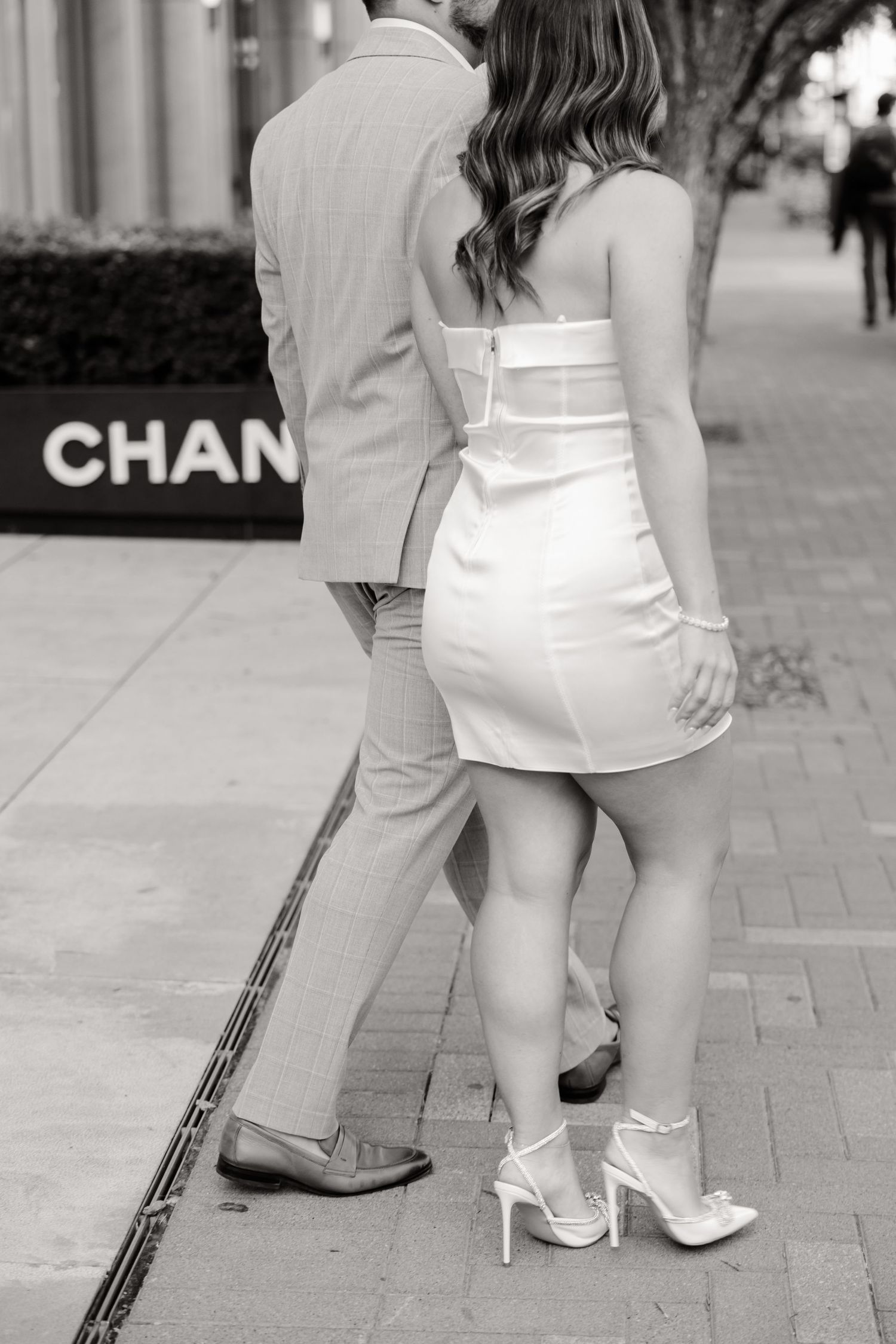 Close-up of a couple's formal attire and footwear outside Chanel, showing white dress and gray suit details.