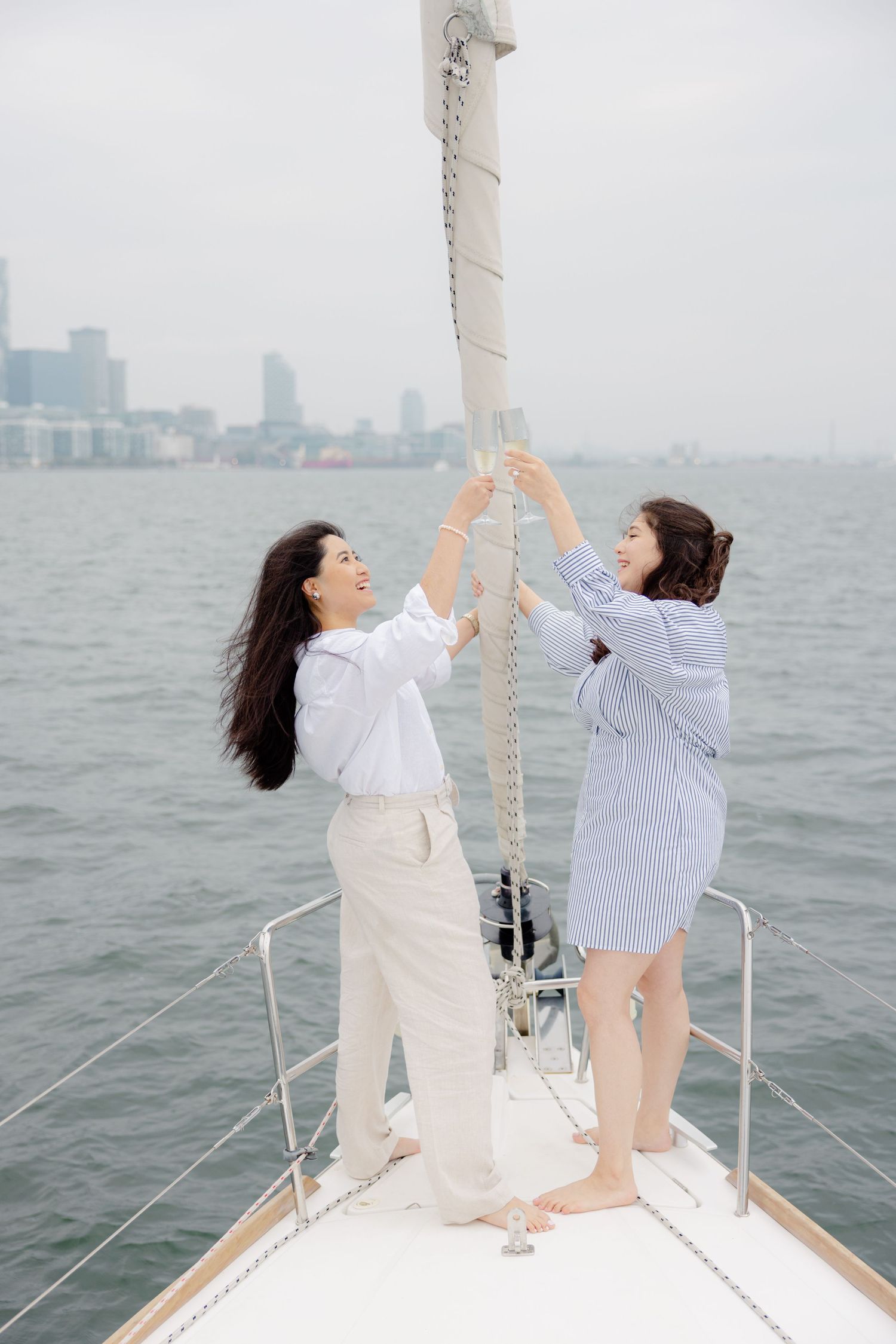 Two people on the bow of a sailing boat pull on ropes against a cityscape backdrop across the water.
