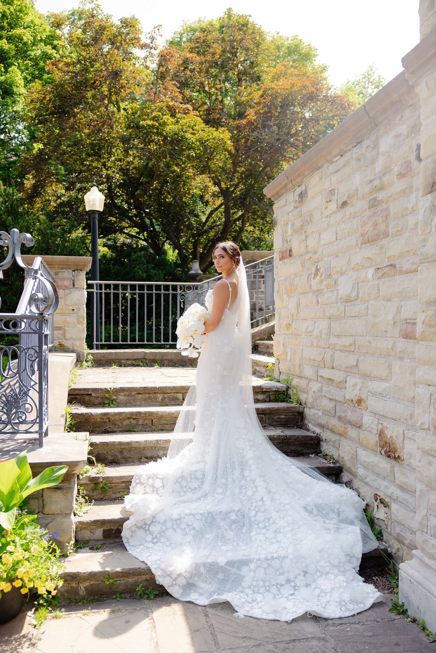 Elegant lace wedding dress with long flowing train cascades down stone garden steps near a decorative iron railing and stone wall.