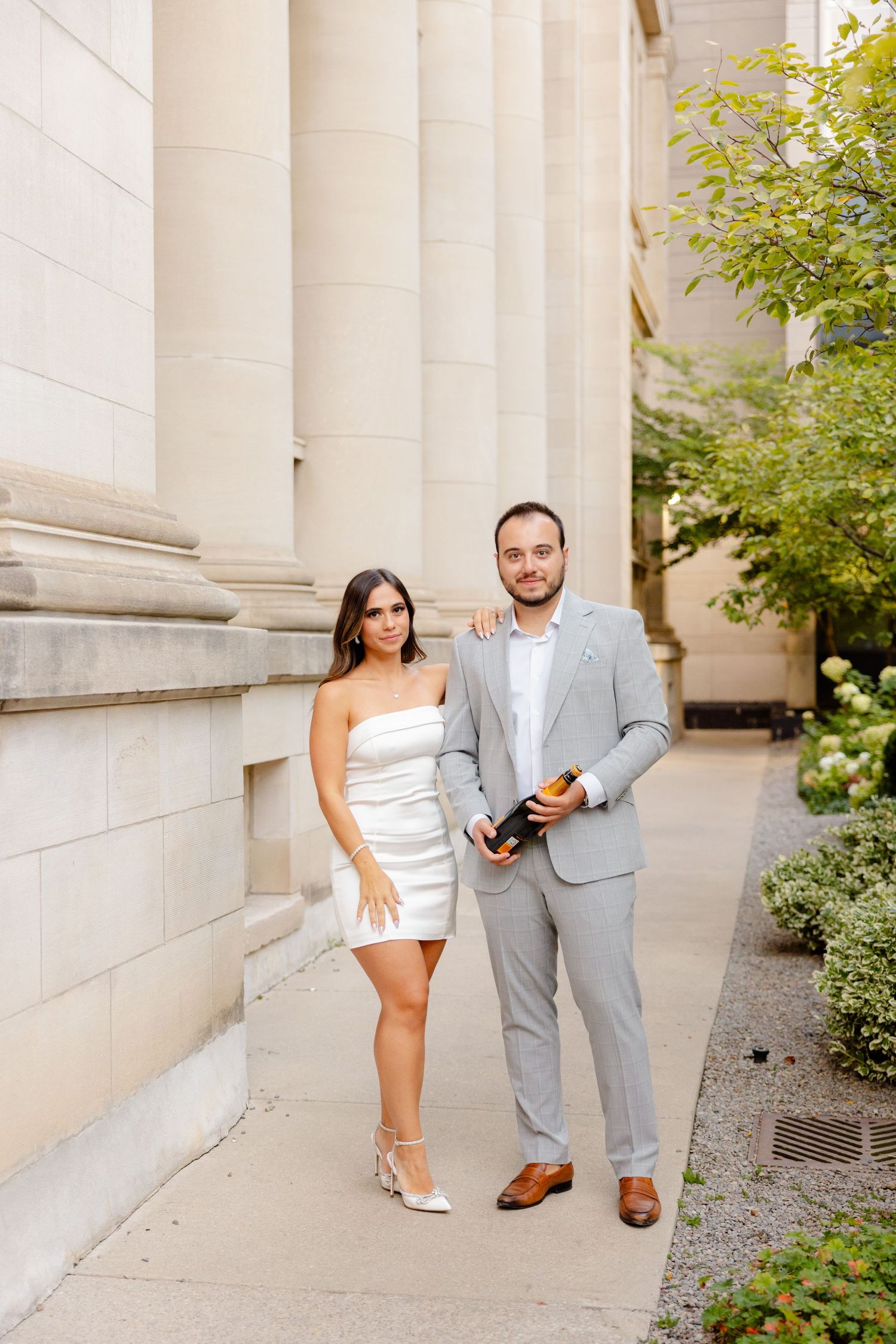 A couple in formal attire poses against white columns, with the woman in white and man in gray holding champagne.