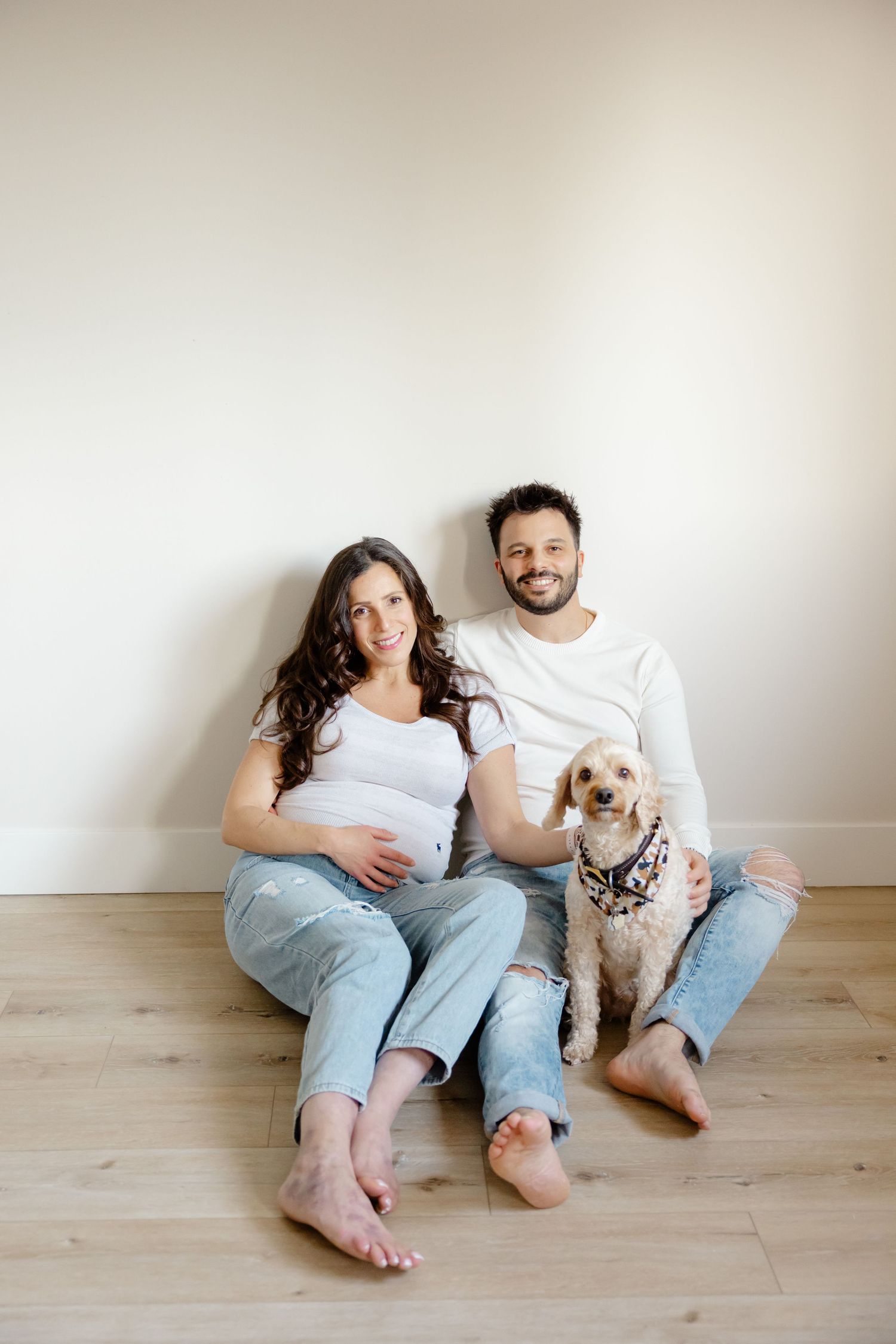 A couple sits on a hardwood floor against a white wall with their small dog, dressed casually in jeans and white tops.