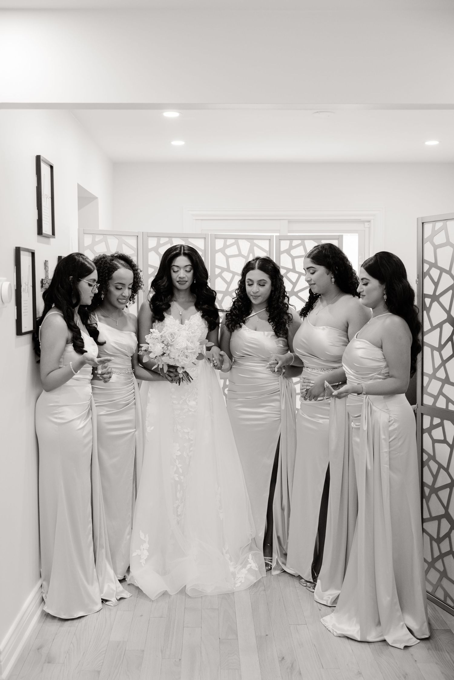 Elegant bridal party in matching floor-length white dresses pose together in a bright indoor setting.