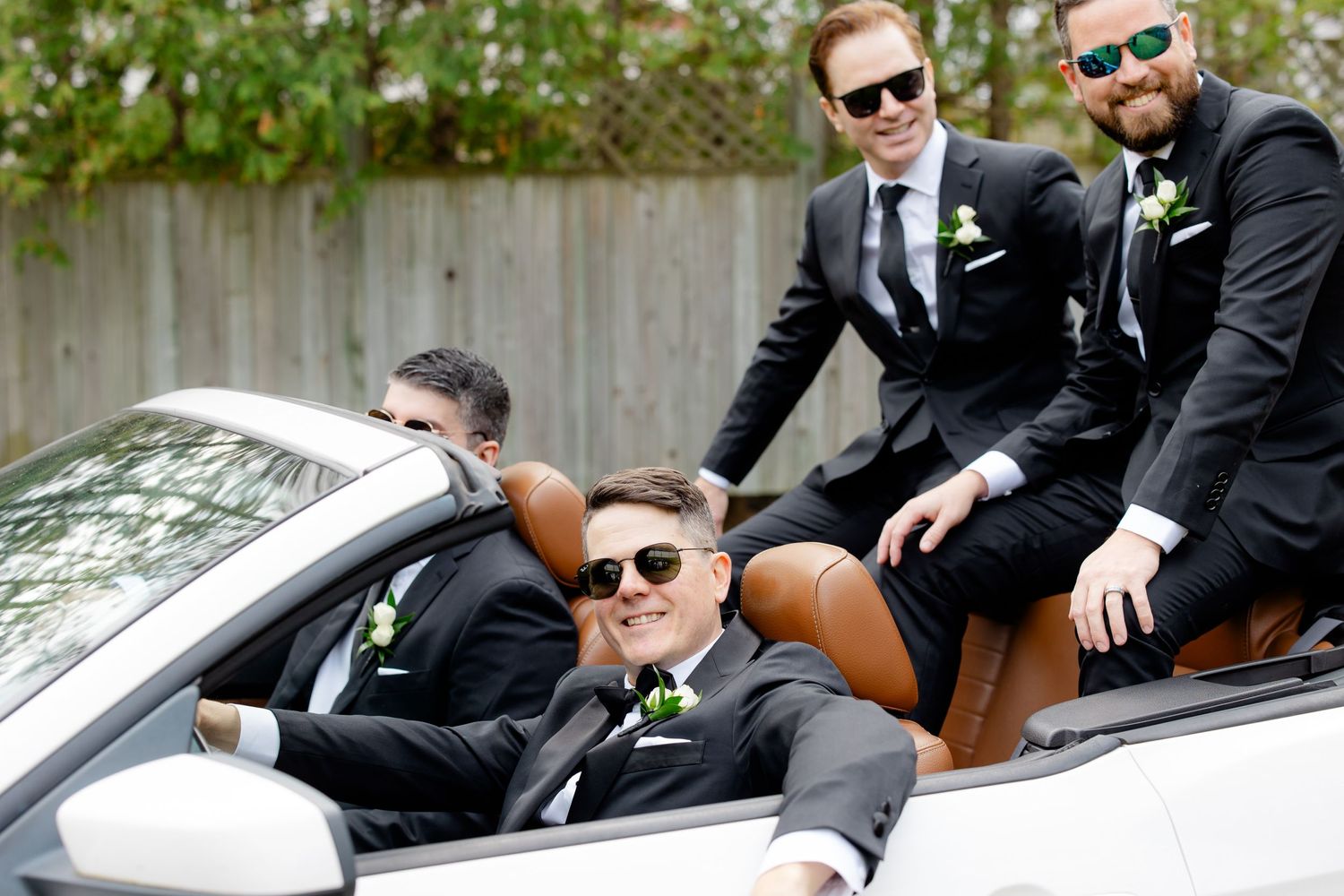Four people in formal attire and sunglasses pose together in and around a white convertible car.