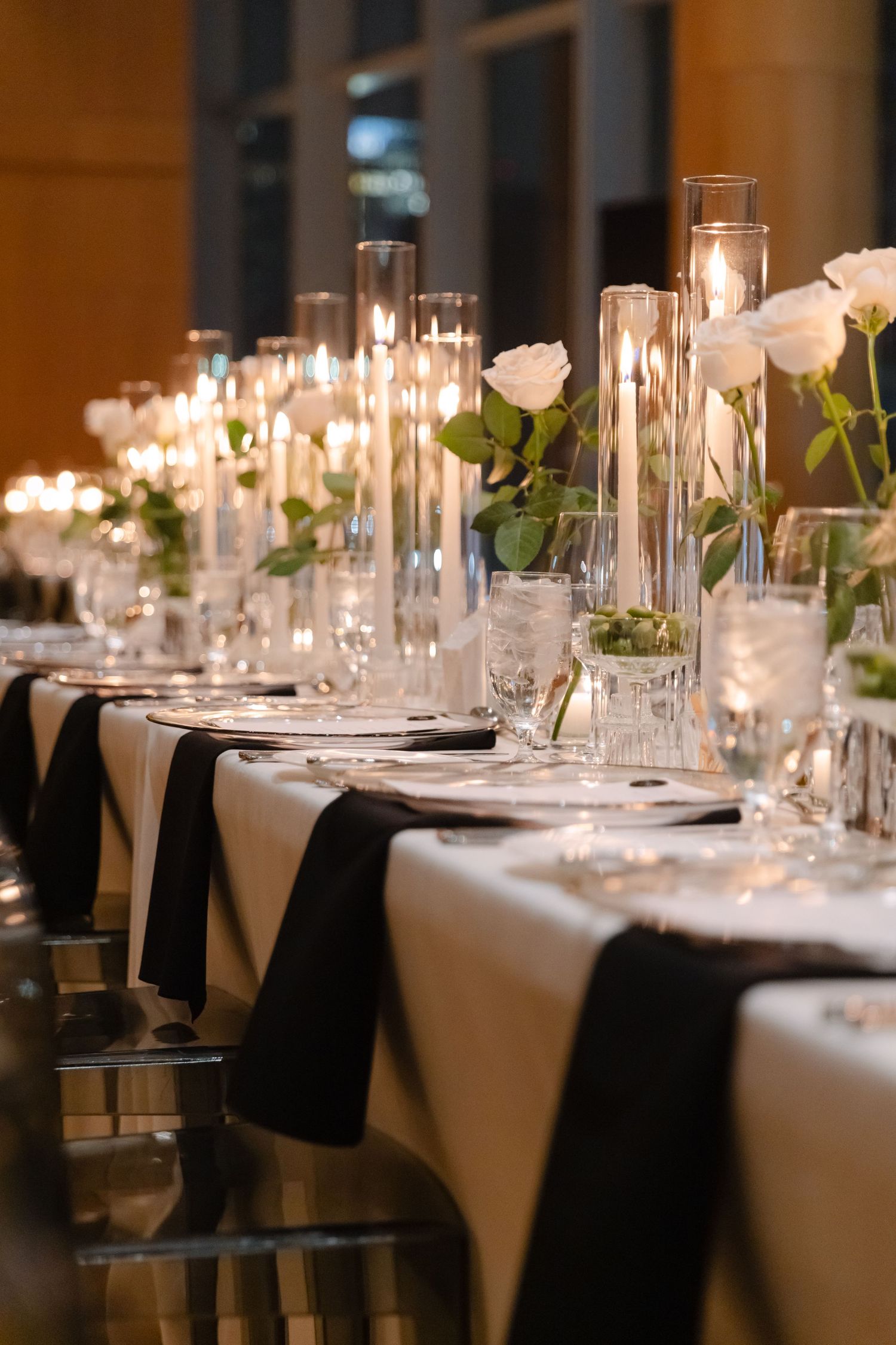 Elegant wedding table setting with white roses, tall glass candleholders, and black napkins on white tablecloth.