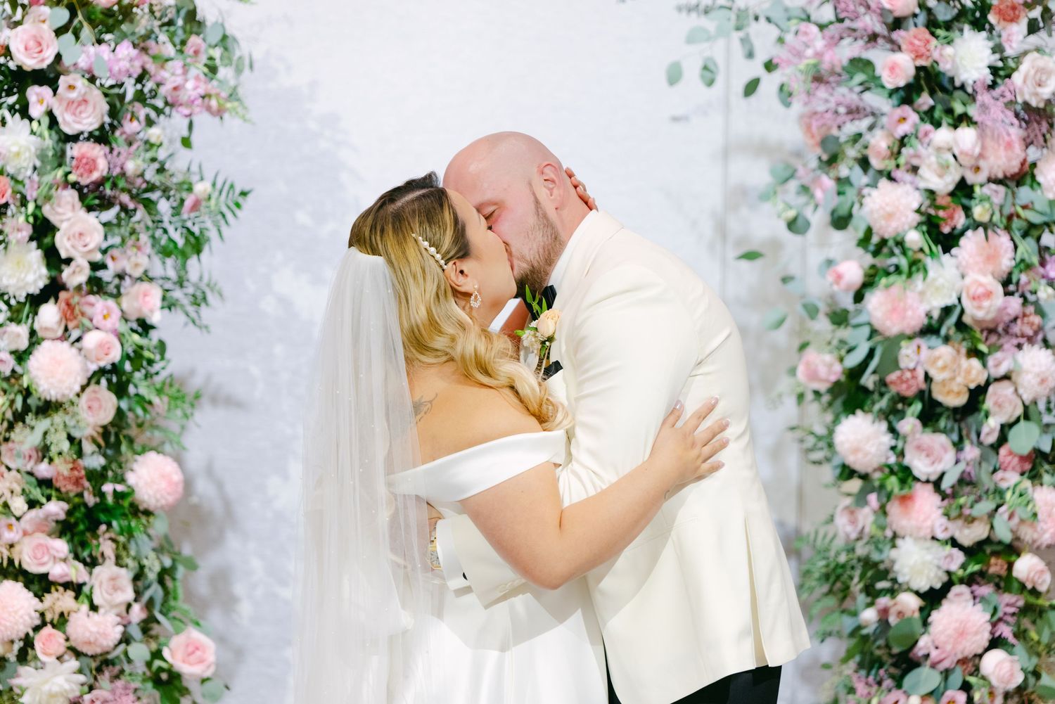 Romantic wedding kiss under beautiful pink rose and greenery arch.