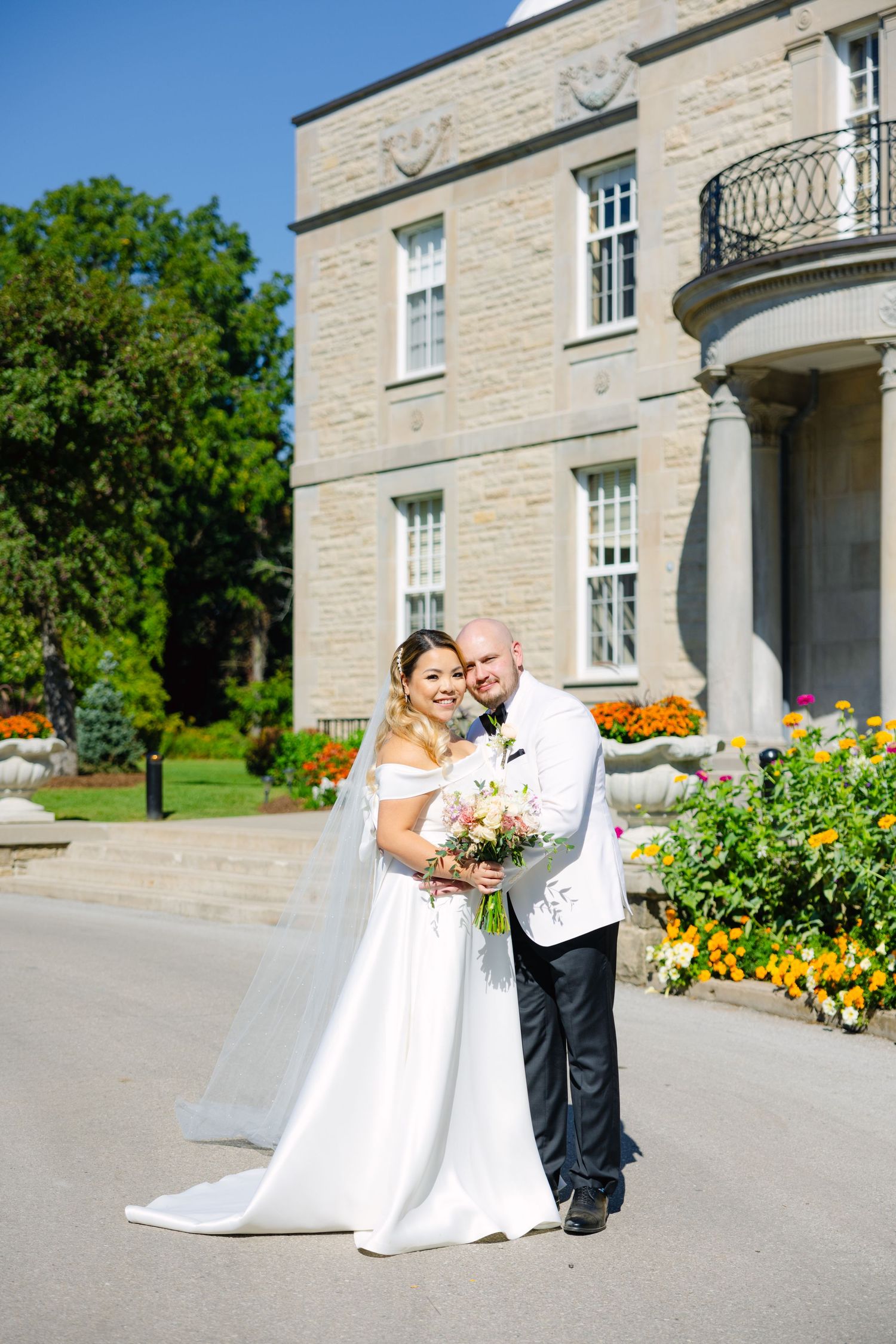 A wedding portrait captured in front of a stately stone building with blooming garden flowers.