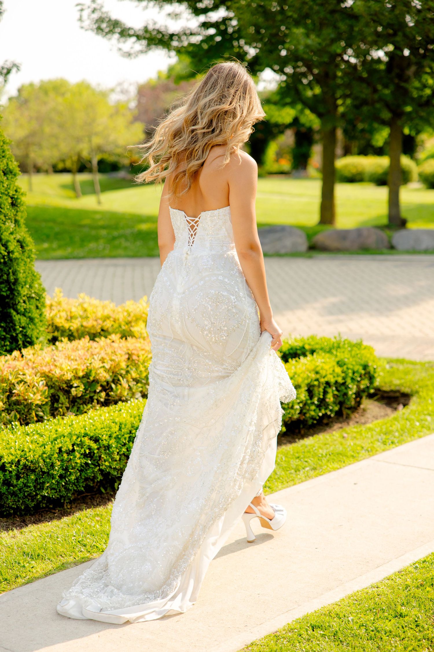 A bride in a flowing lace wedding gown walks along a garden path surrounded by manicured hedges and trees.