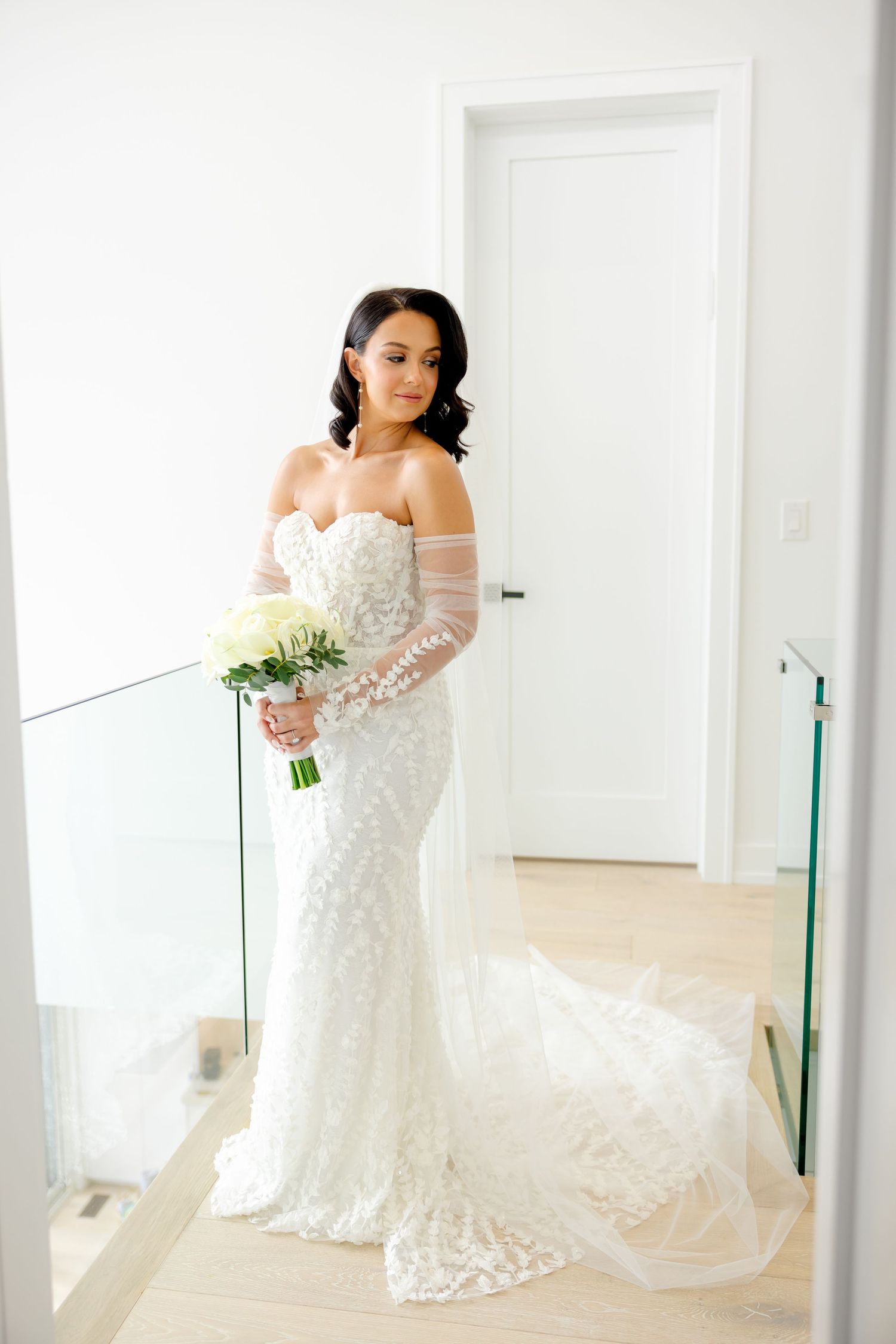 Full-length bridal portrait showcasing intricate lace dress and cream bouquet against white interior.