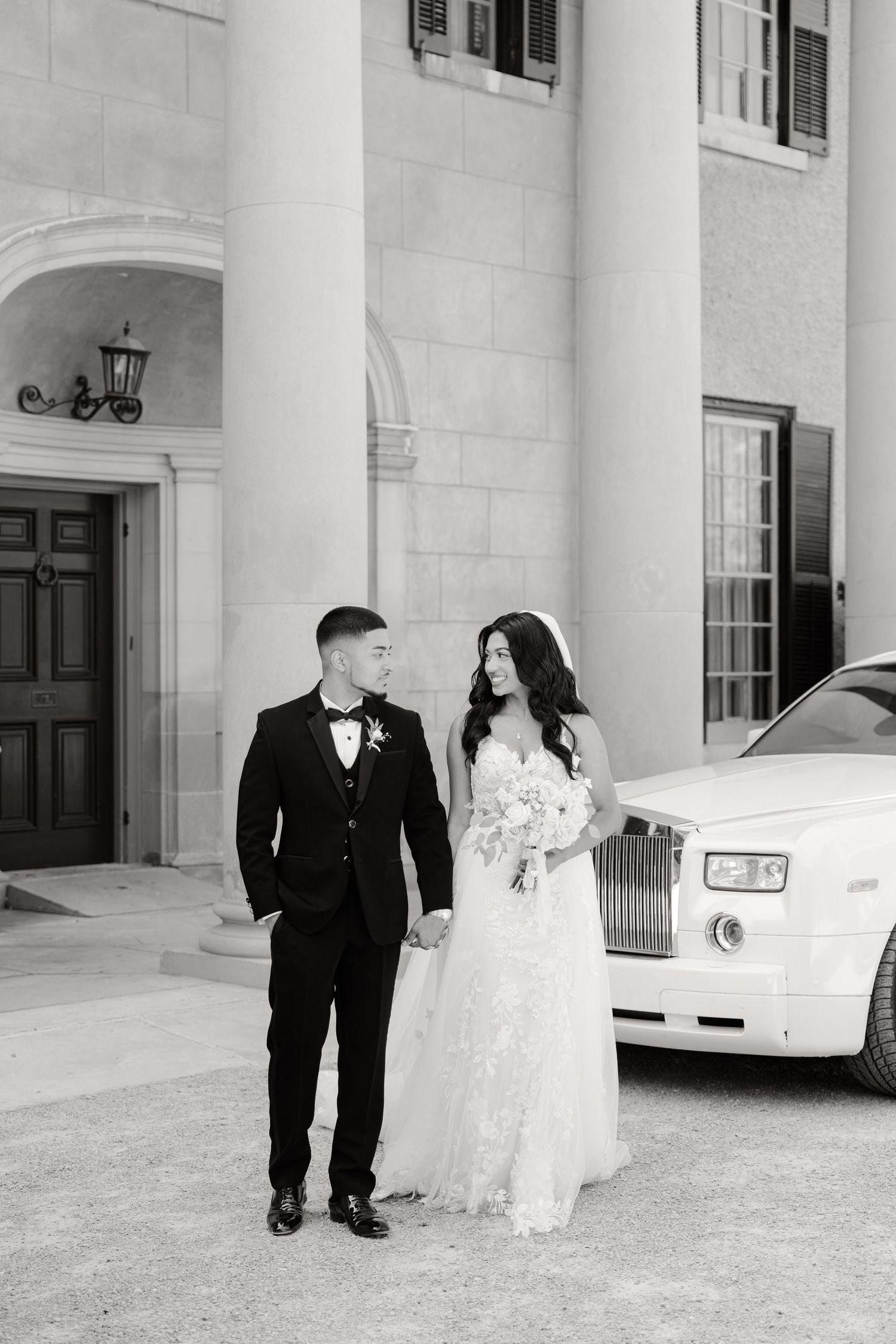 Black and white wedding portrait of a couple walking together near a luxury car.