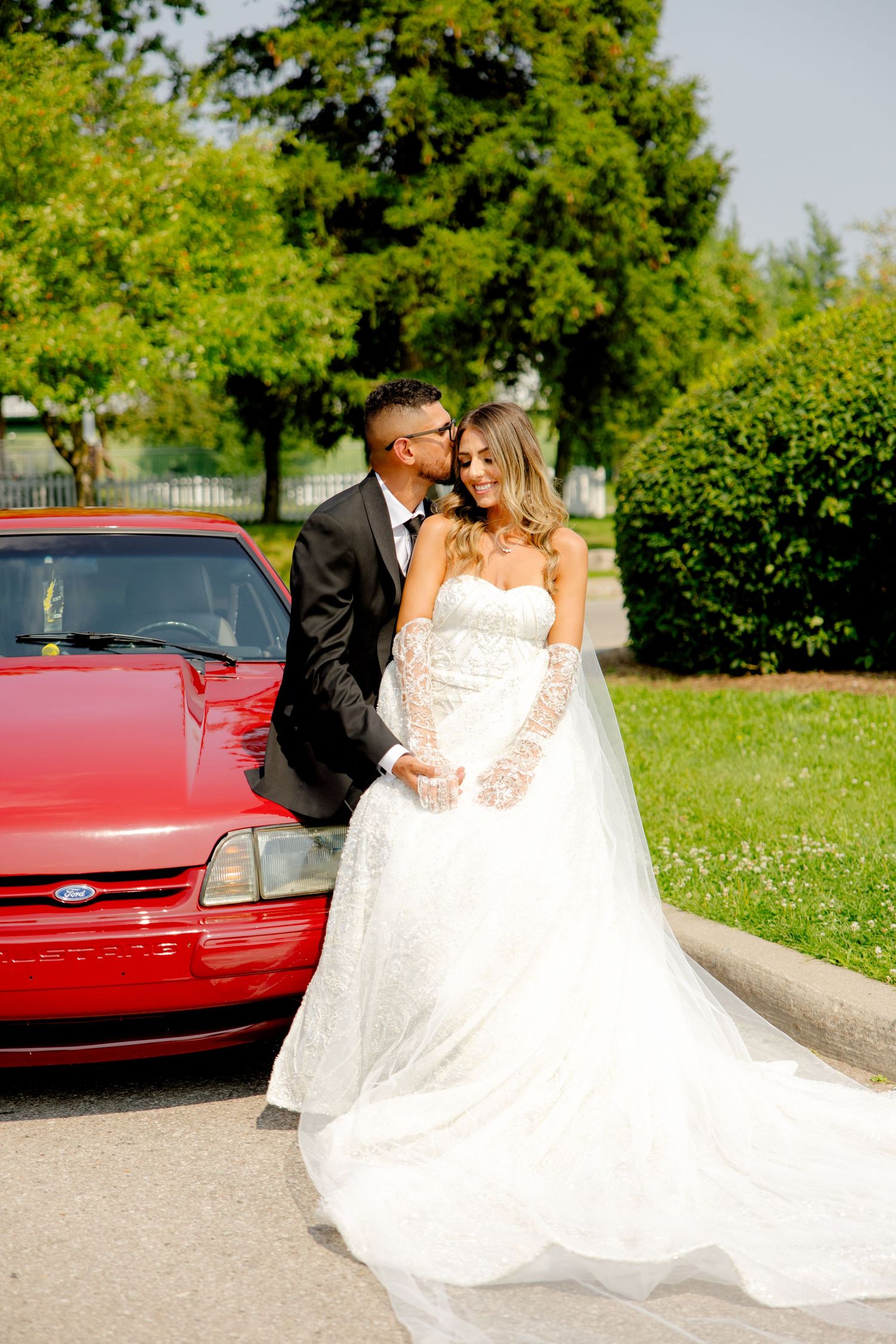 Wedding couple shares an intimate moment beside a bright red classic car surrounded by lush greenery.