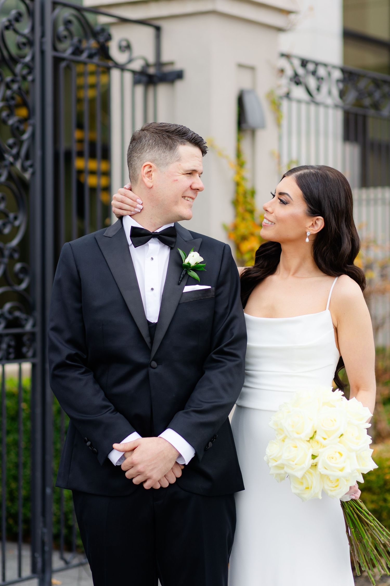 Elegant couple in formal wedding attire share a moment in front of ornate black gates.