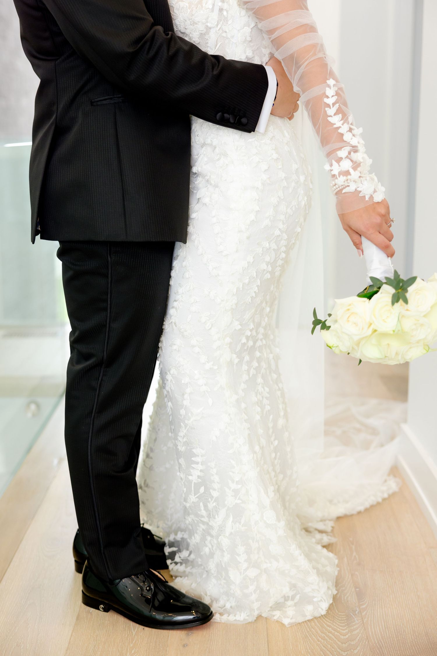 Detail shot of bride's lace gown and groom's formal attire as they stand close together.