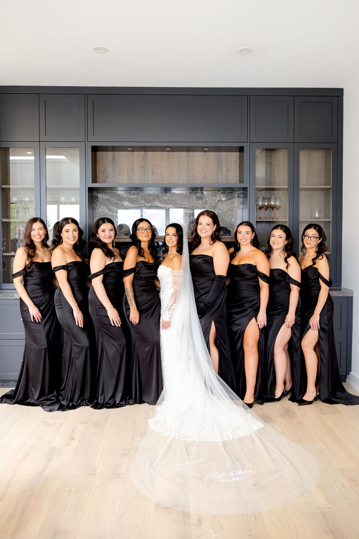 Wedding party lineup featuring bridesmaids in black dresses with thigh slits beside bride in white gown.