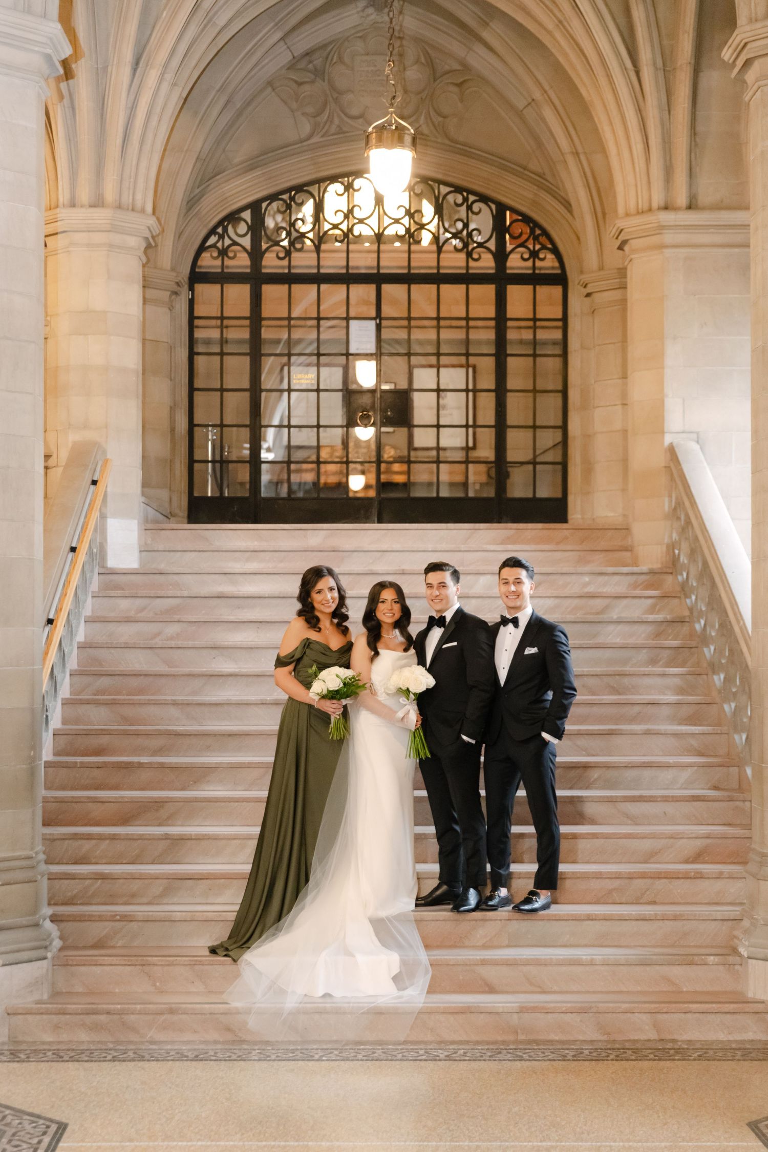 Bridal party stands together on grand marble steps beneath architectural arched window with decorative ironwork.