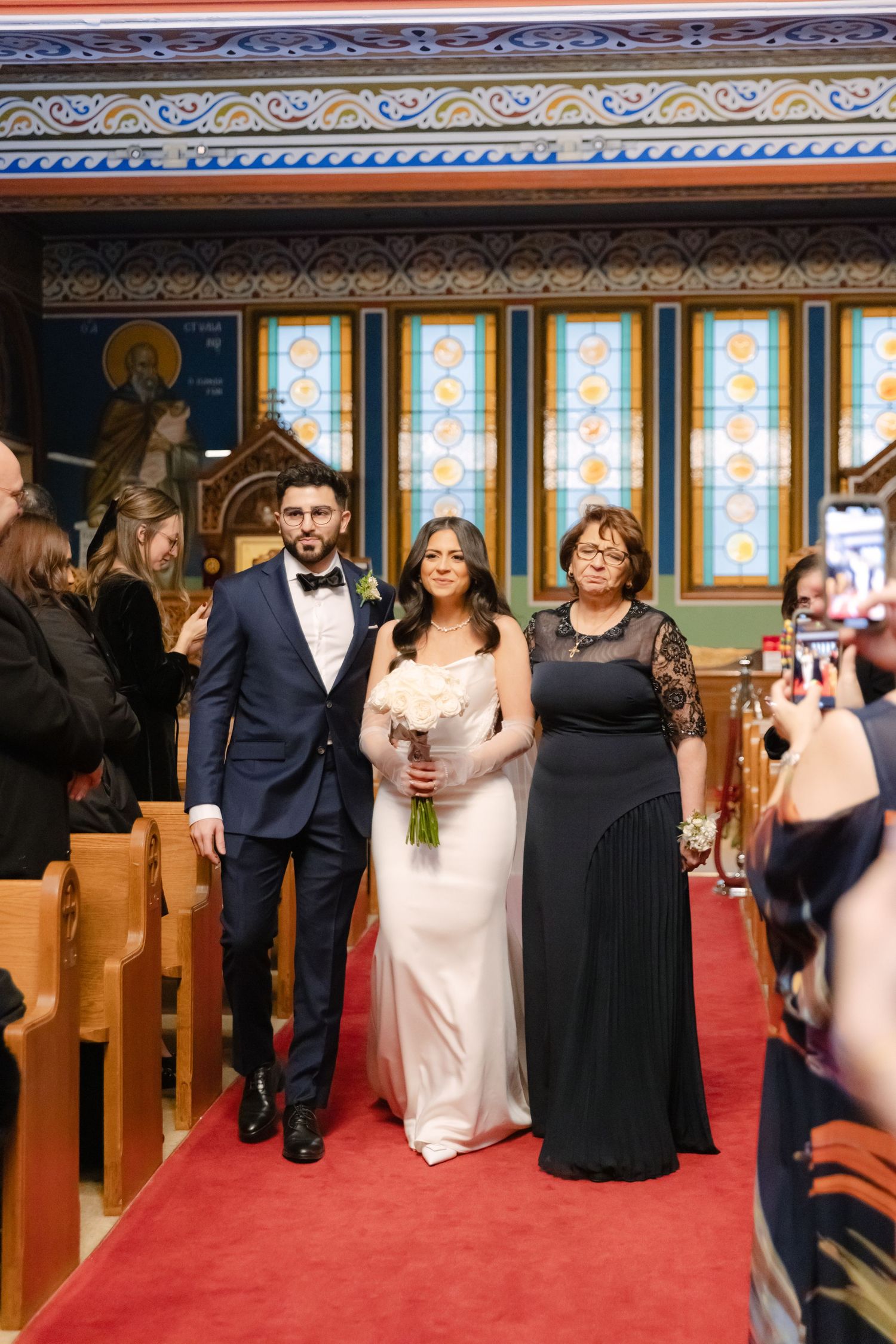 Wedding party walks down red-carpeted church aisle under ornate ceiling with stained glass windows.