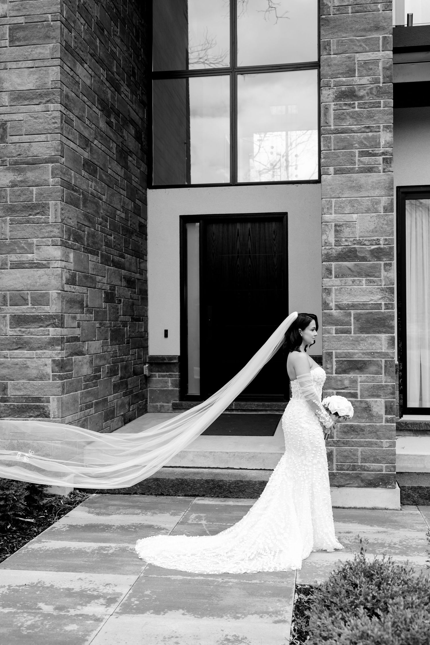 Black and white photo of a bride with flowing veil against stone building backdrop.