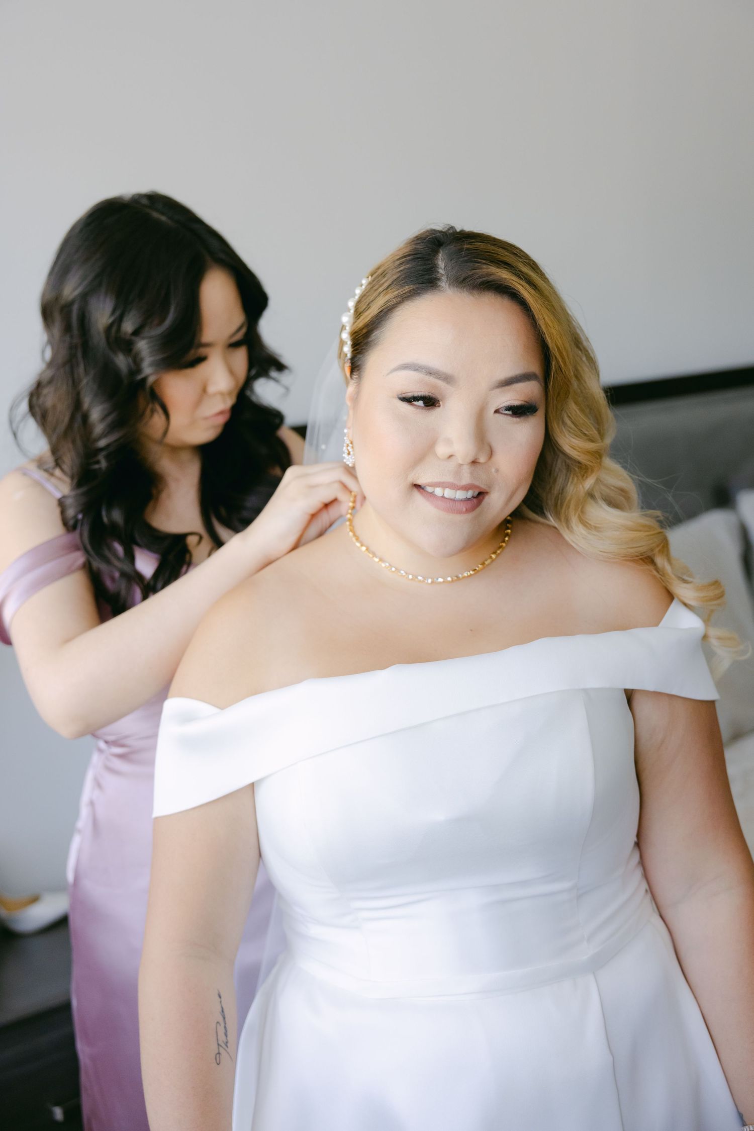 Two people preparing for a wedding, with one helping adjust the bride's off-shoulder white gown.