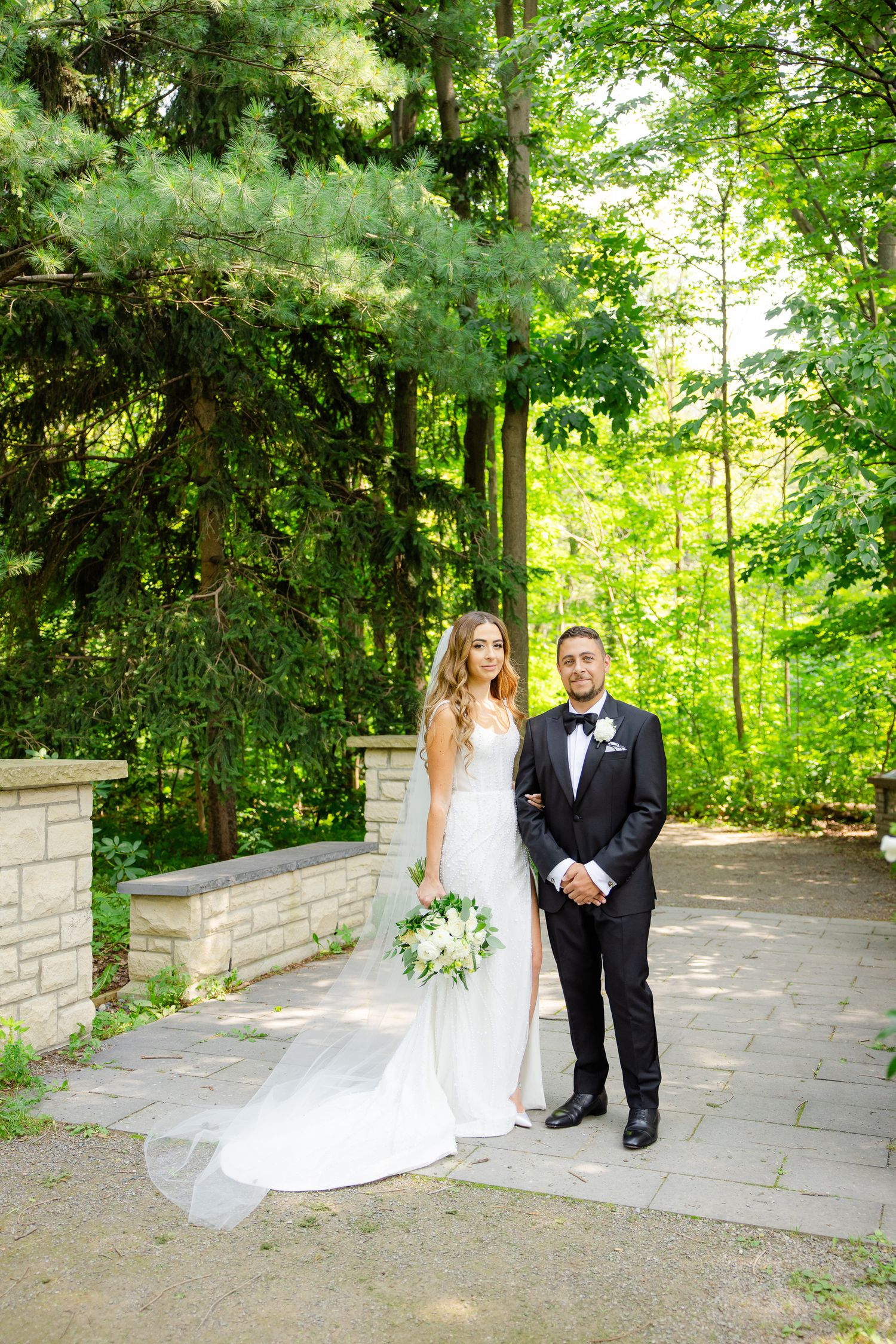 Newlyweds pose on stone pathway surrounded by lush green trees and landscaping.
