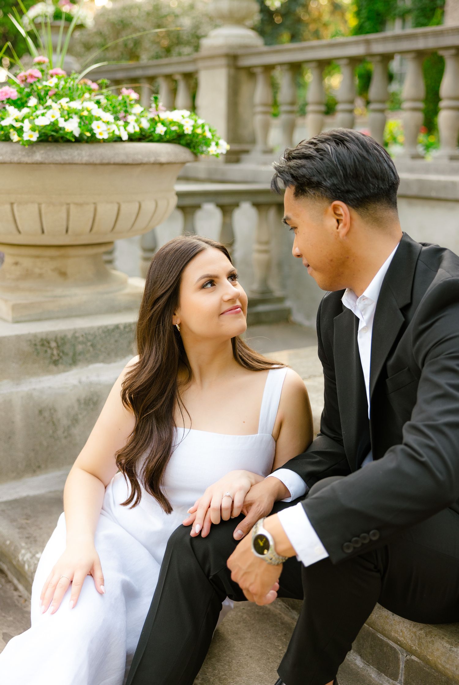 A couple in formal wear shares a tender moment on stone steps with flowering planters in the background.