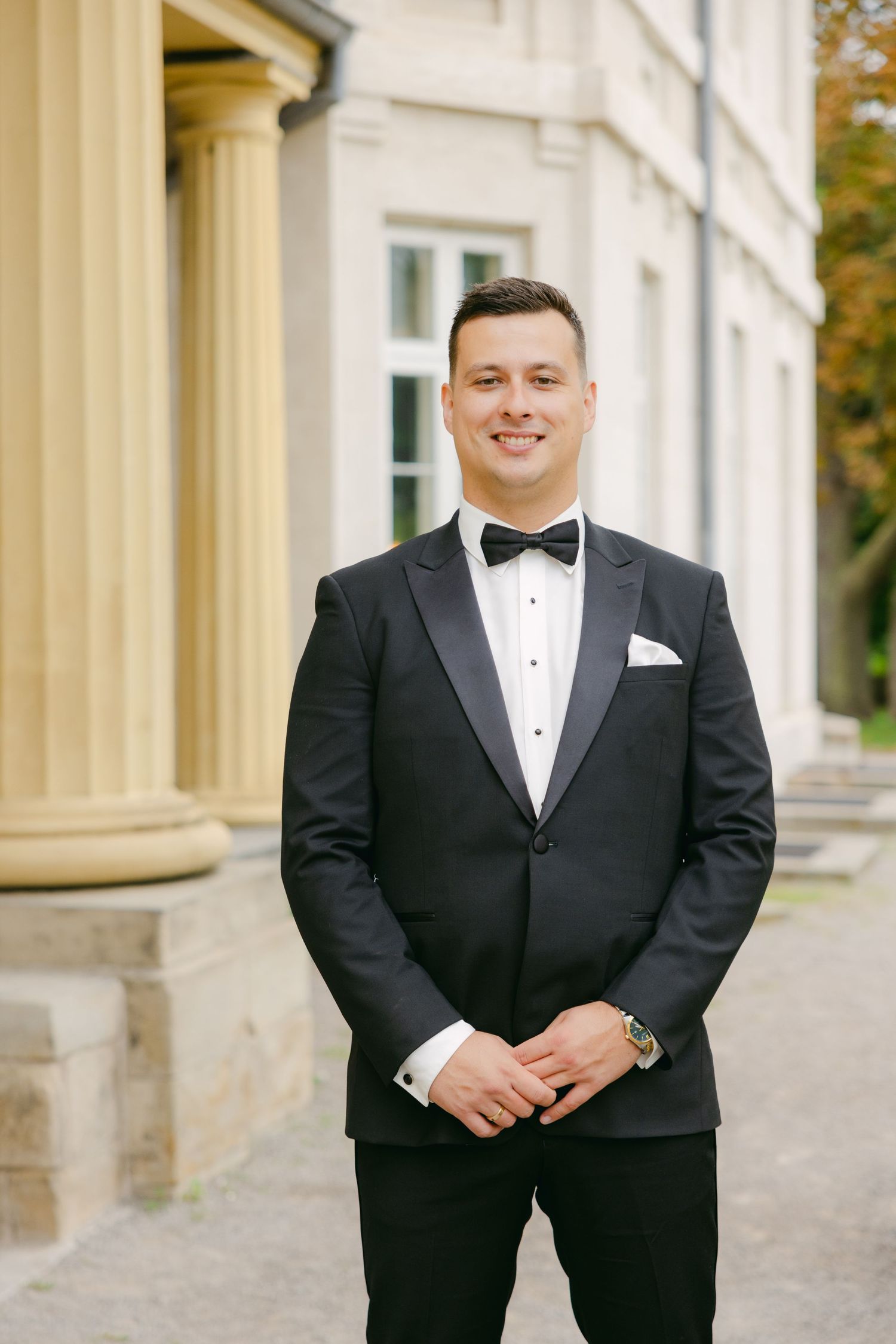 Groom in black tuxedo smiles while standing beside classical architecture.