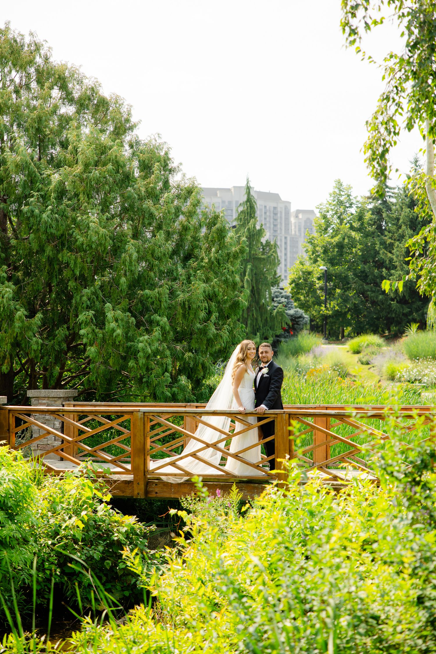 Newlyweds pose on a wooden bridge in a scenic garden with city buildings visible in background.
