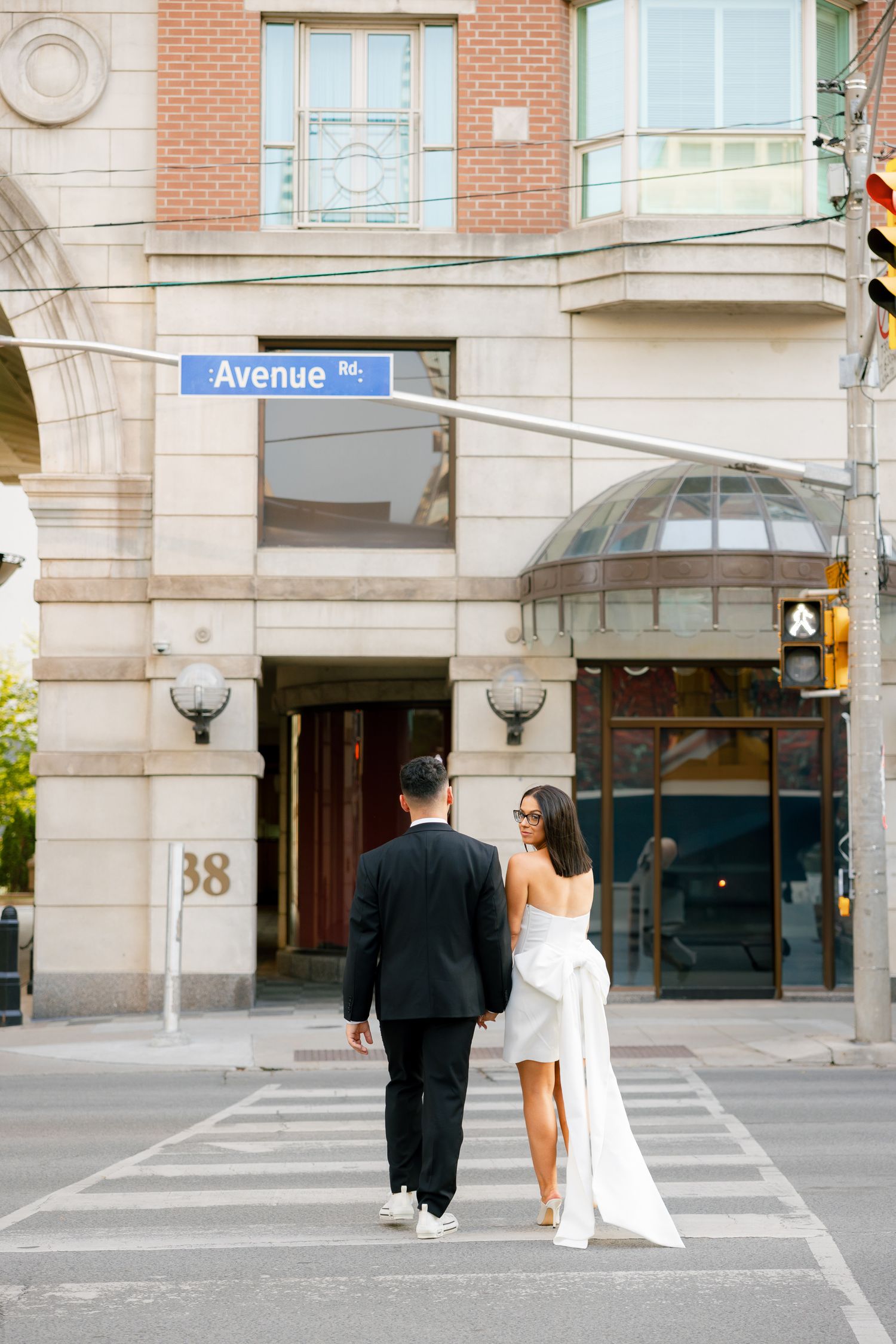 A couple in wedding attire walking hand in hand across a city crosswalk near Avenue sign.