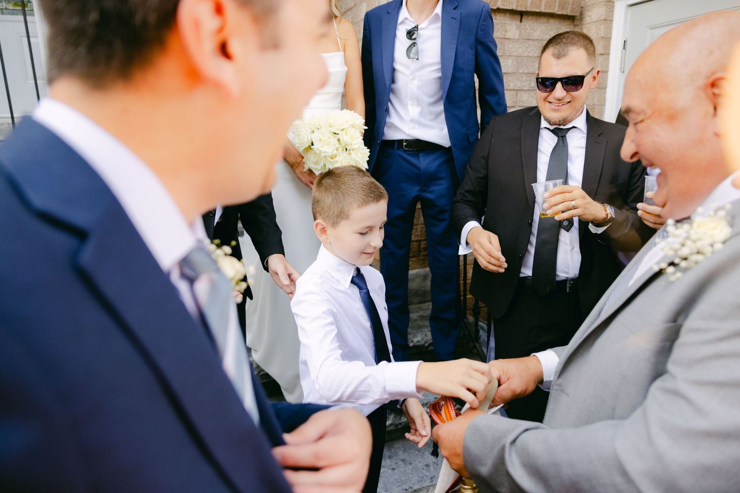 Wedding guests in formal attire gather outside while young child in white shirt mingles.