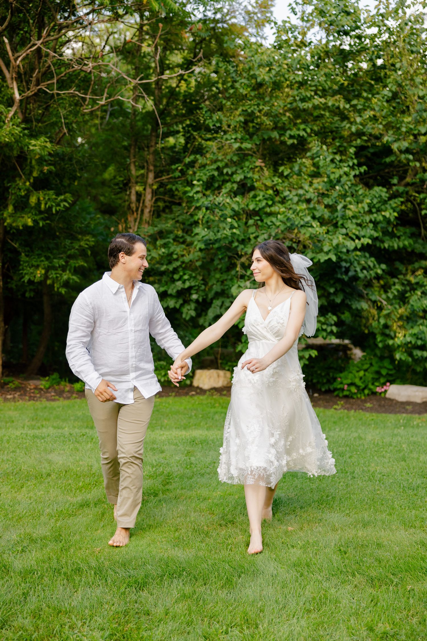 A carefree couple walking barefoot on grass in wedding attire.