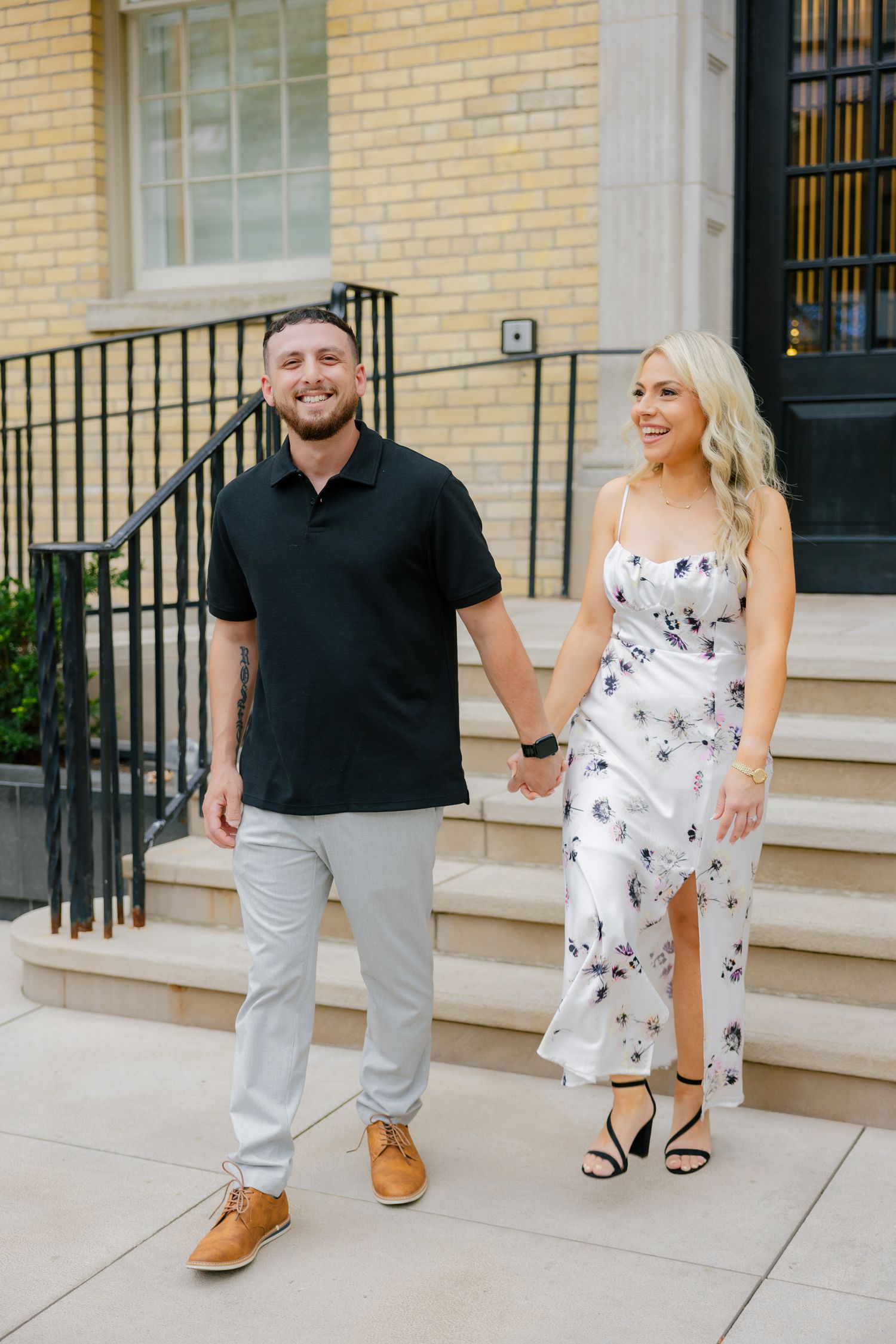 A couple stands holding hands on stone steps in front of a yellow brick building, wearing contrasting black and floral outfits.
