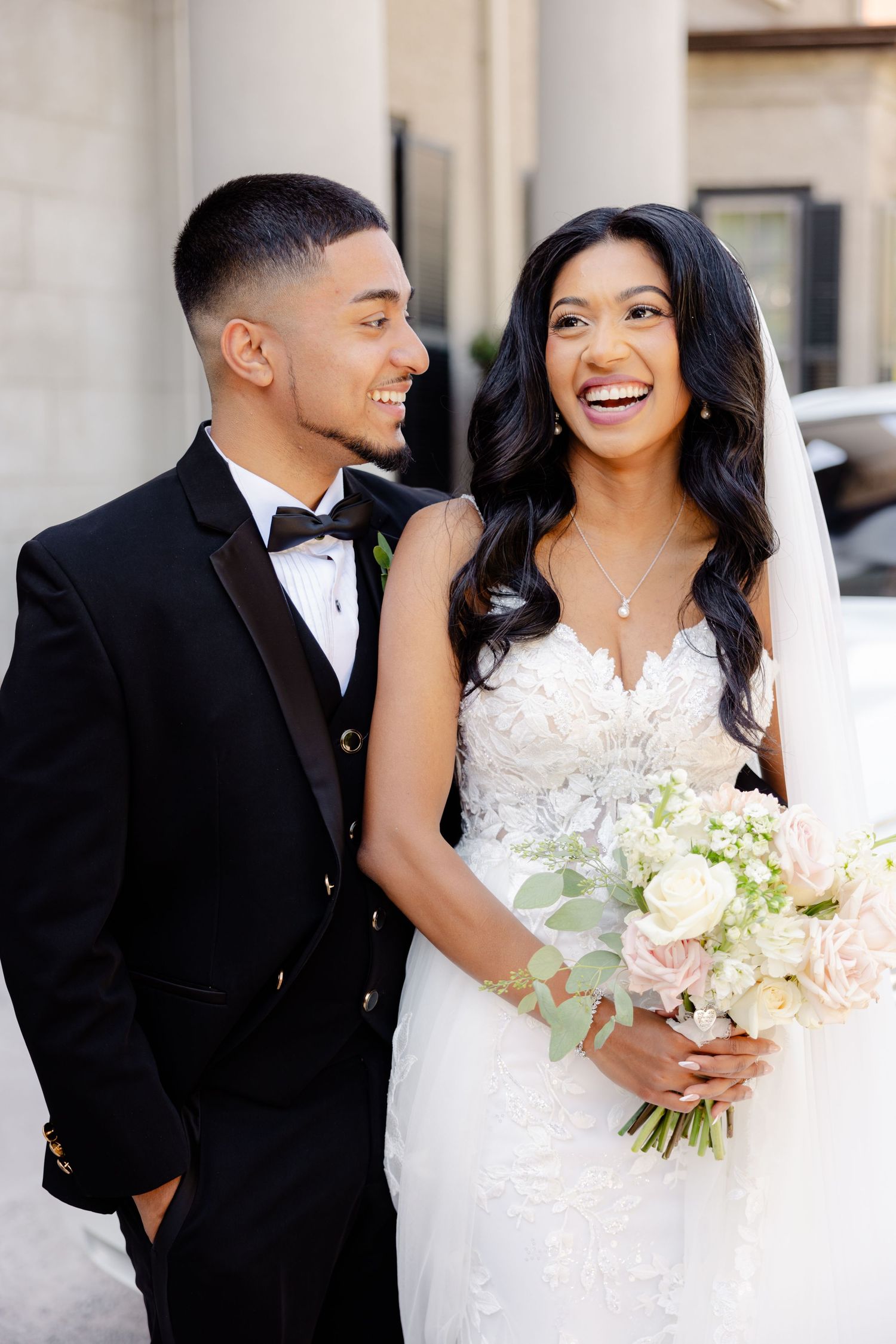 A wedding couple shares a joyful laugh together while wearing formal attire and holding a bouquet of white flowers.