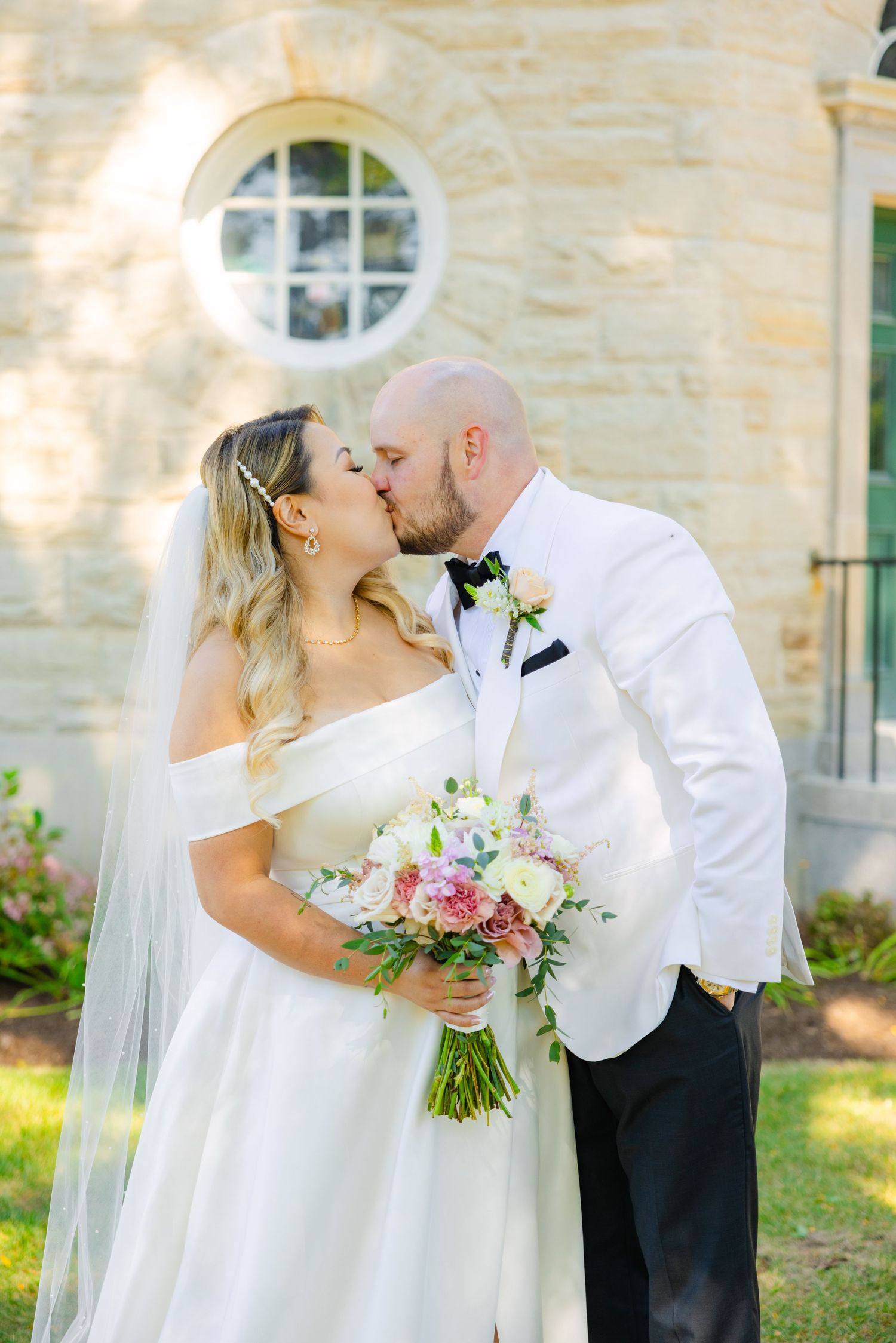 The newlyweds share a joyful kiss while holding a pink and white bouquet in front of stone architecture.