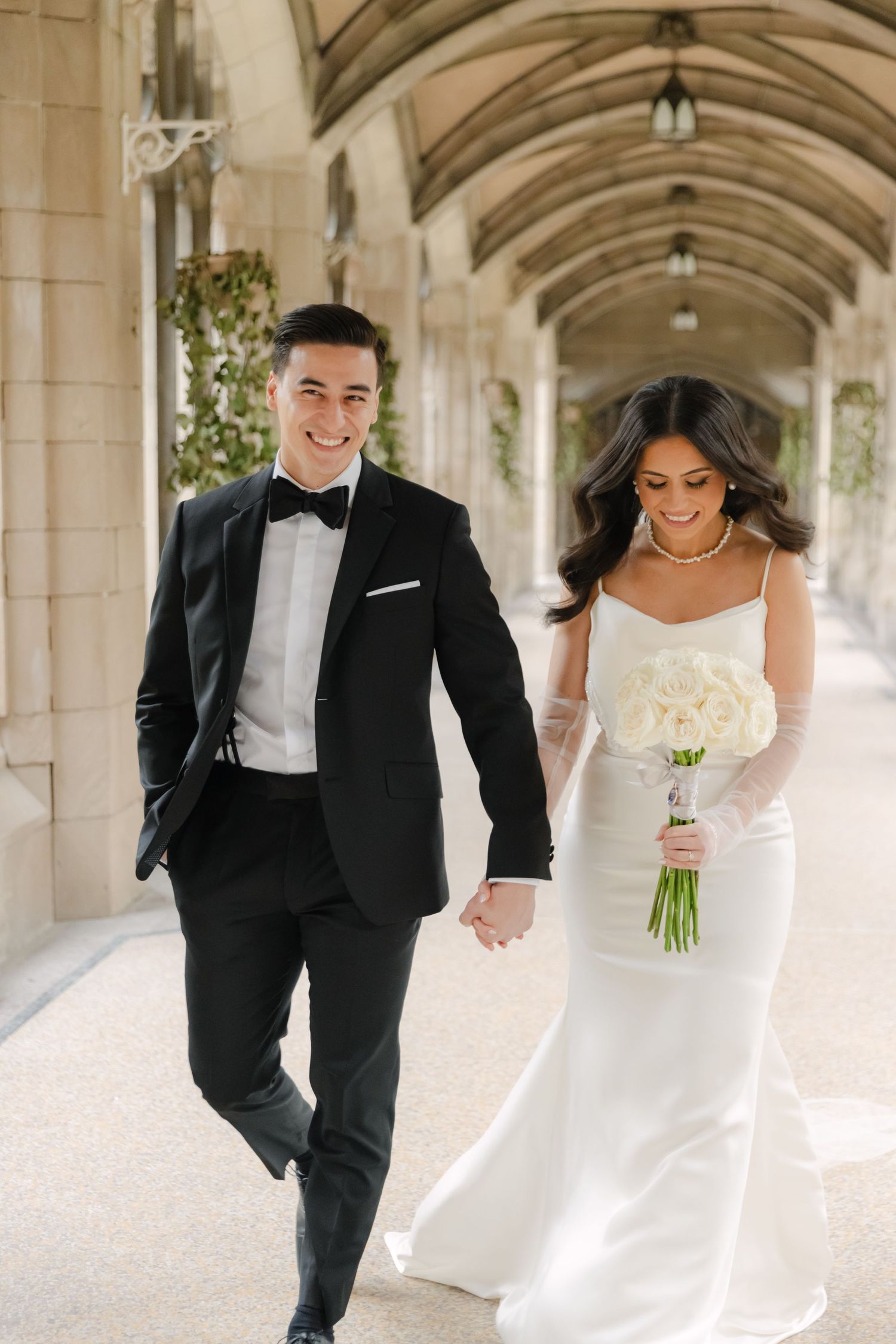 A bride in white gown holds a bouquet while walking with her groom in a black tuxedo through an elegant arched hallway.