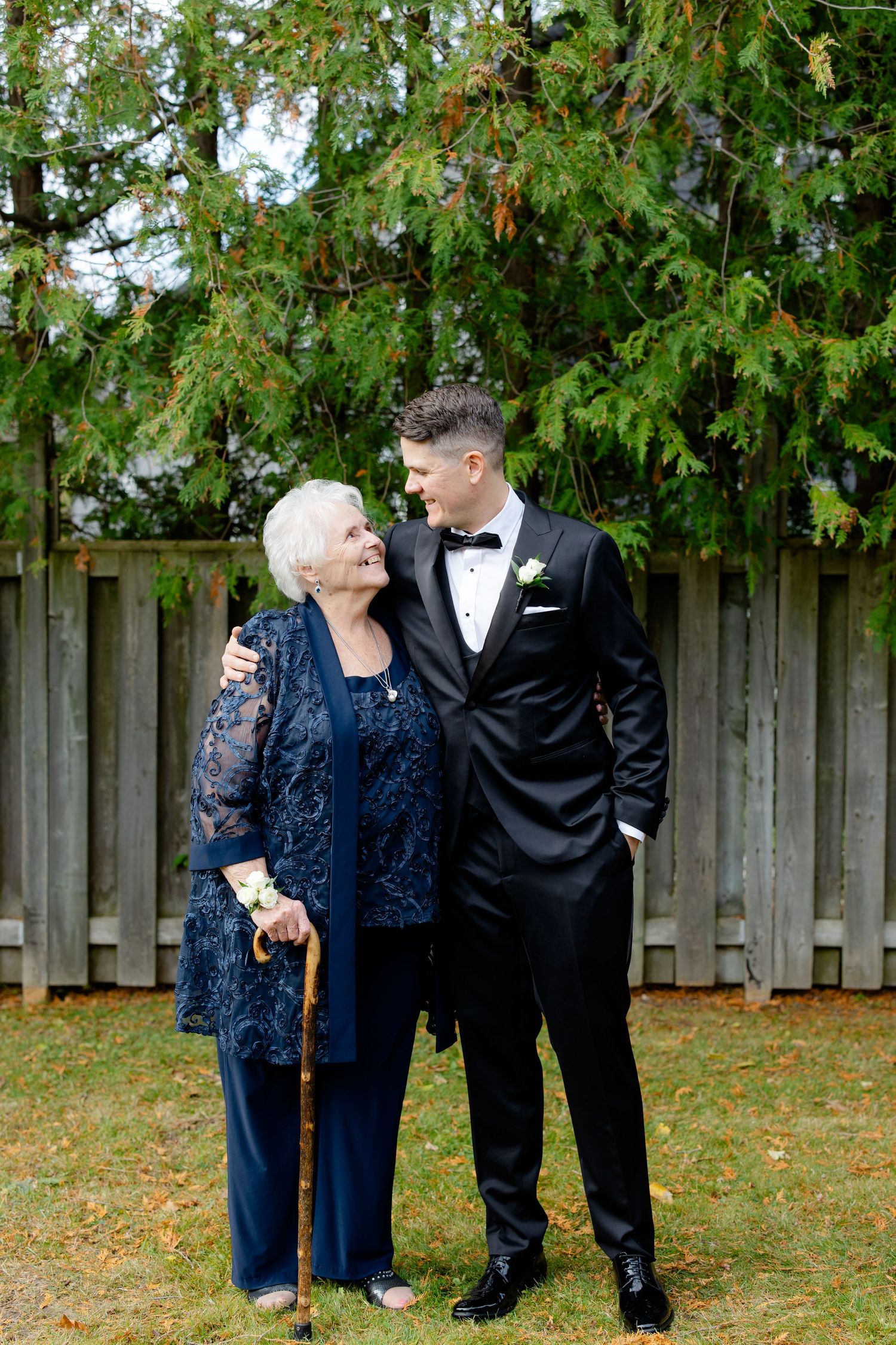 Two elegantly dressed people share a tender moment while standing in front of a wooden fence and greenery.